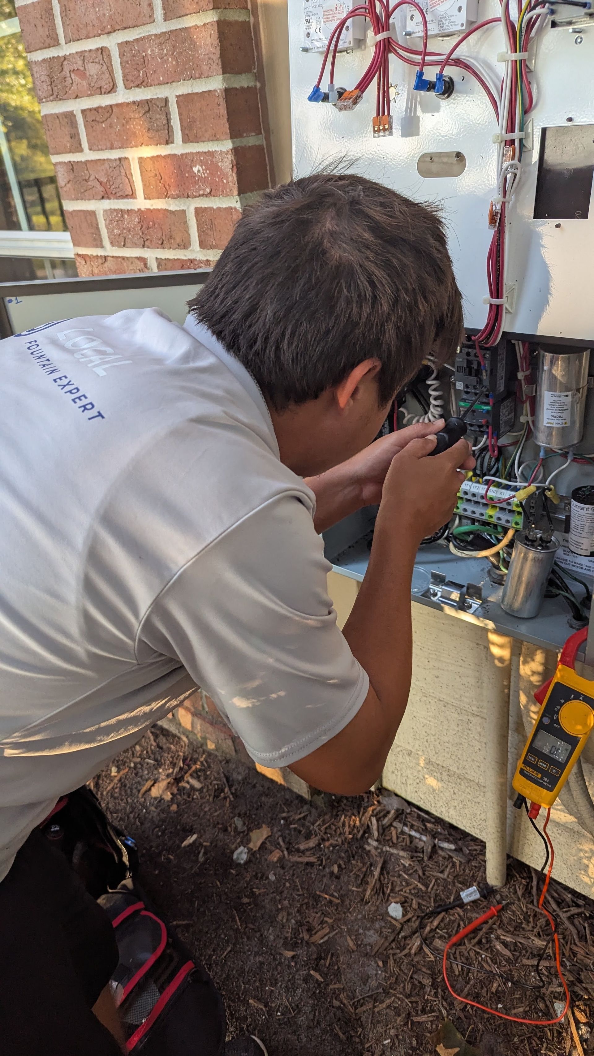 Person repairs electrical equipment outside, holding tools. Multimeter, wires visible.