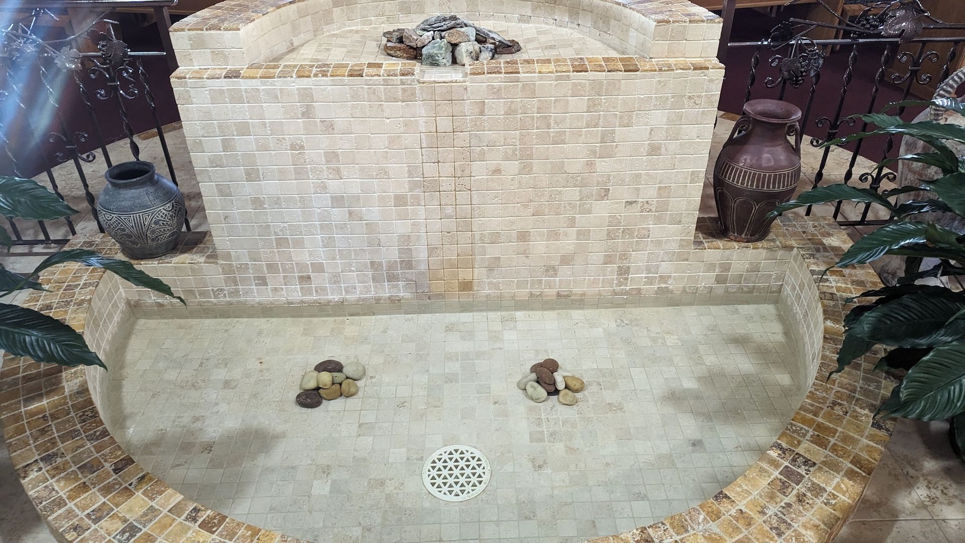 Indoor water fountain with a stone-tiled wall and basin. Features pottery and greenery.