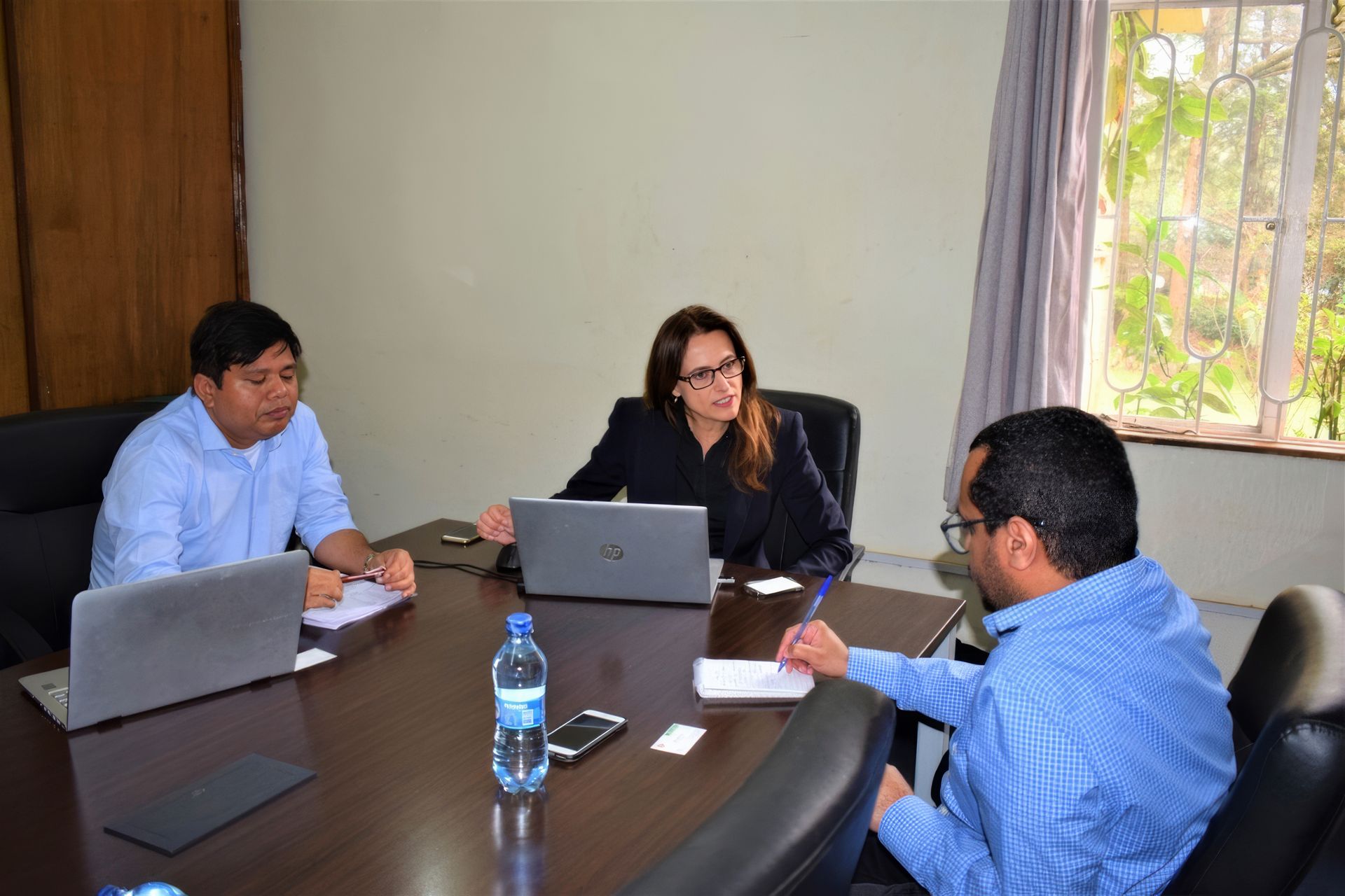 Three people in a meeting around a table with laptops in a bright office.