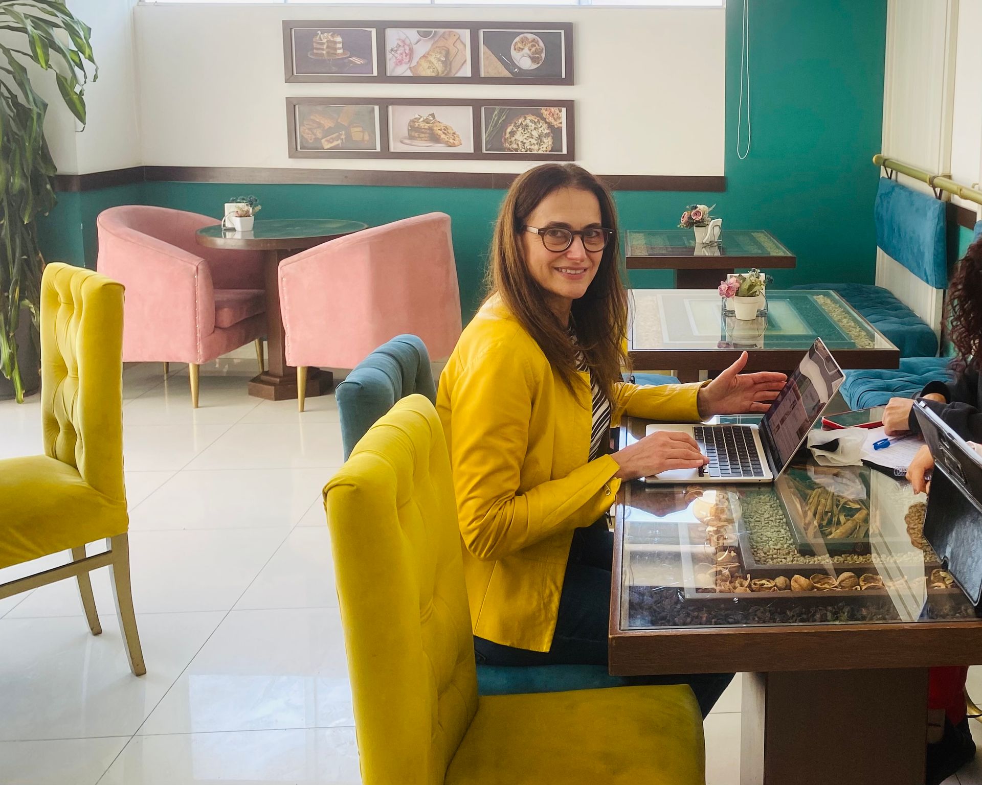 Woman in a yellow blazer smiling at a laptop in a bright café with pink and teal seating.