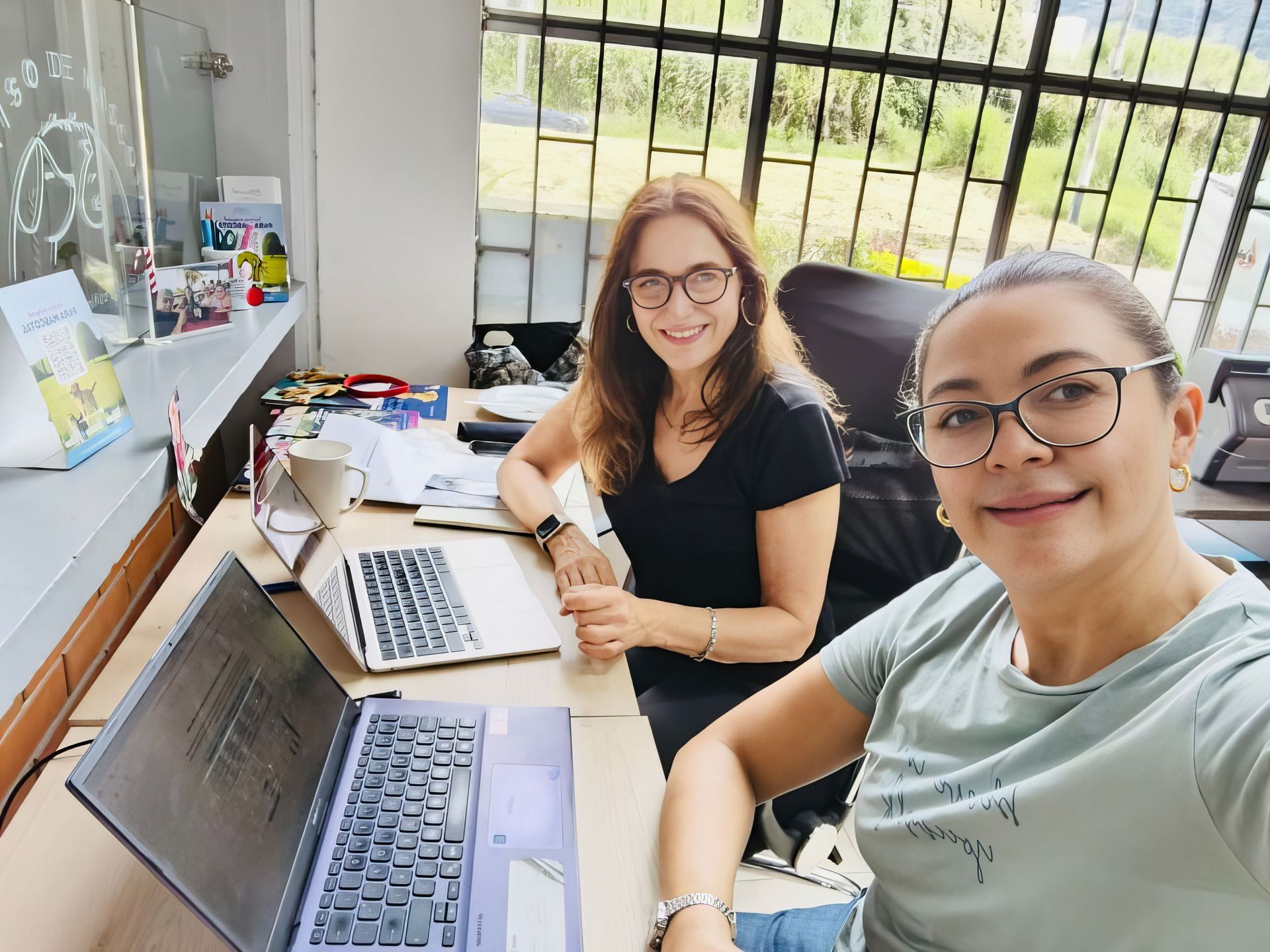 Two people smiling at a desk with open laptops in a bright room by a window