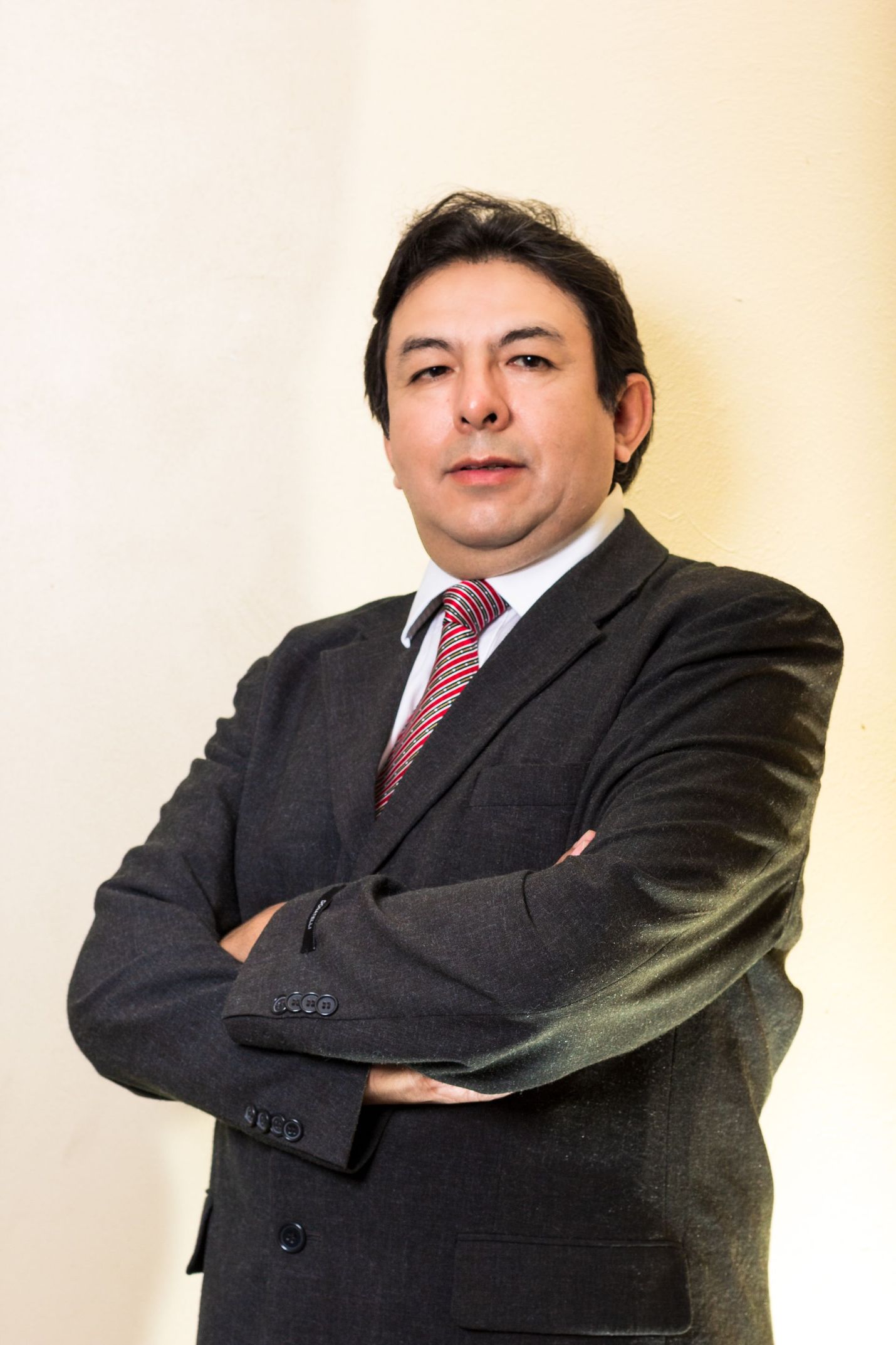 Man in a dark suit and striped tie stands with arms crossed against a light background.