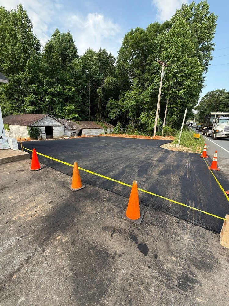 Freshly paved asphalt surface, orange cones, and yellow caution tape. A building and trees are in the background.