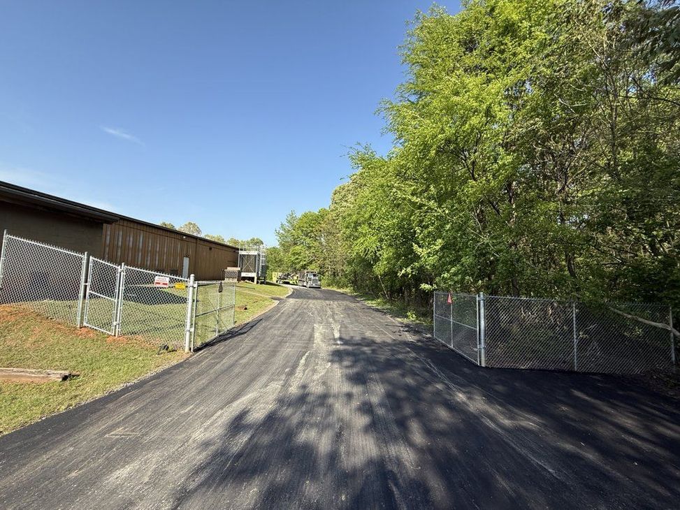 Paved road leading to a warehouse with a chain-link fence and trees on either side under a blue sky.
