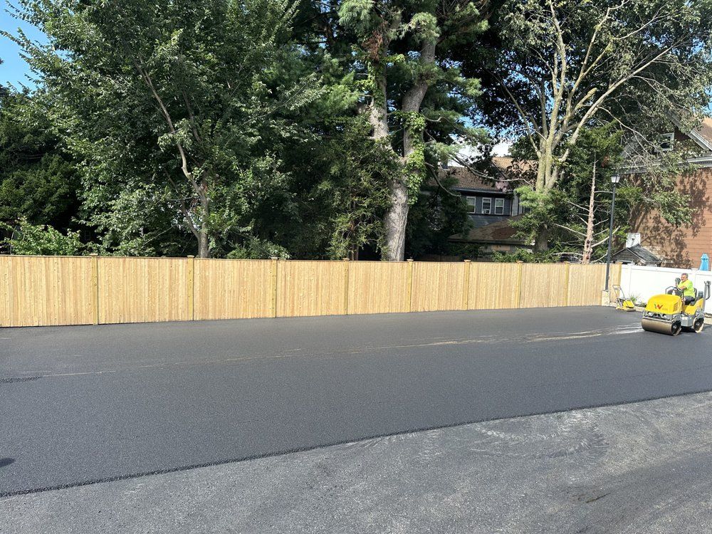 Asphalt paving with a roller in action, against a wooden fence and trees.