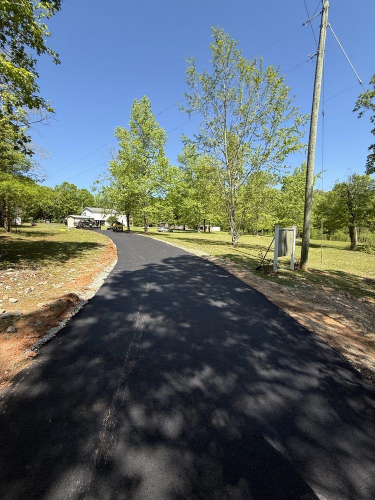 A newly paved asphalt driveway winds towards a house, surrounded by green trees and grass on a sunny day.