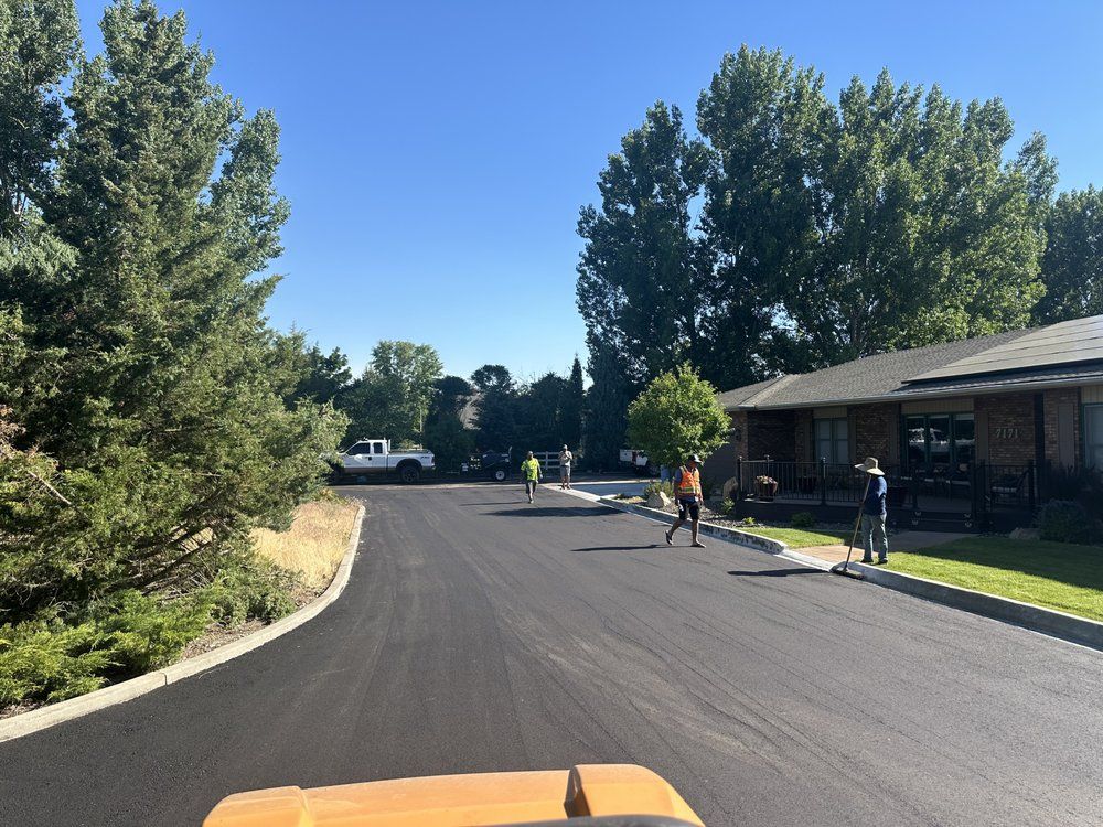 Paved road with construction workers; bright sun, trees and a house in the background.