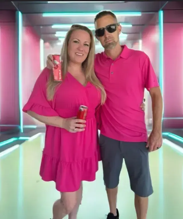 Couple in pink outfits holding drinks, smiling in front of a futuristic, pink-lit background.