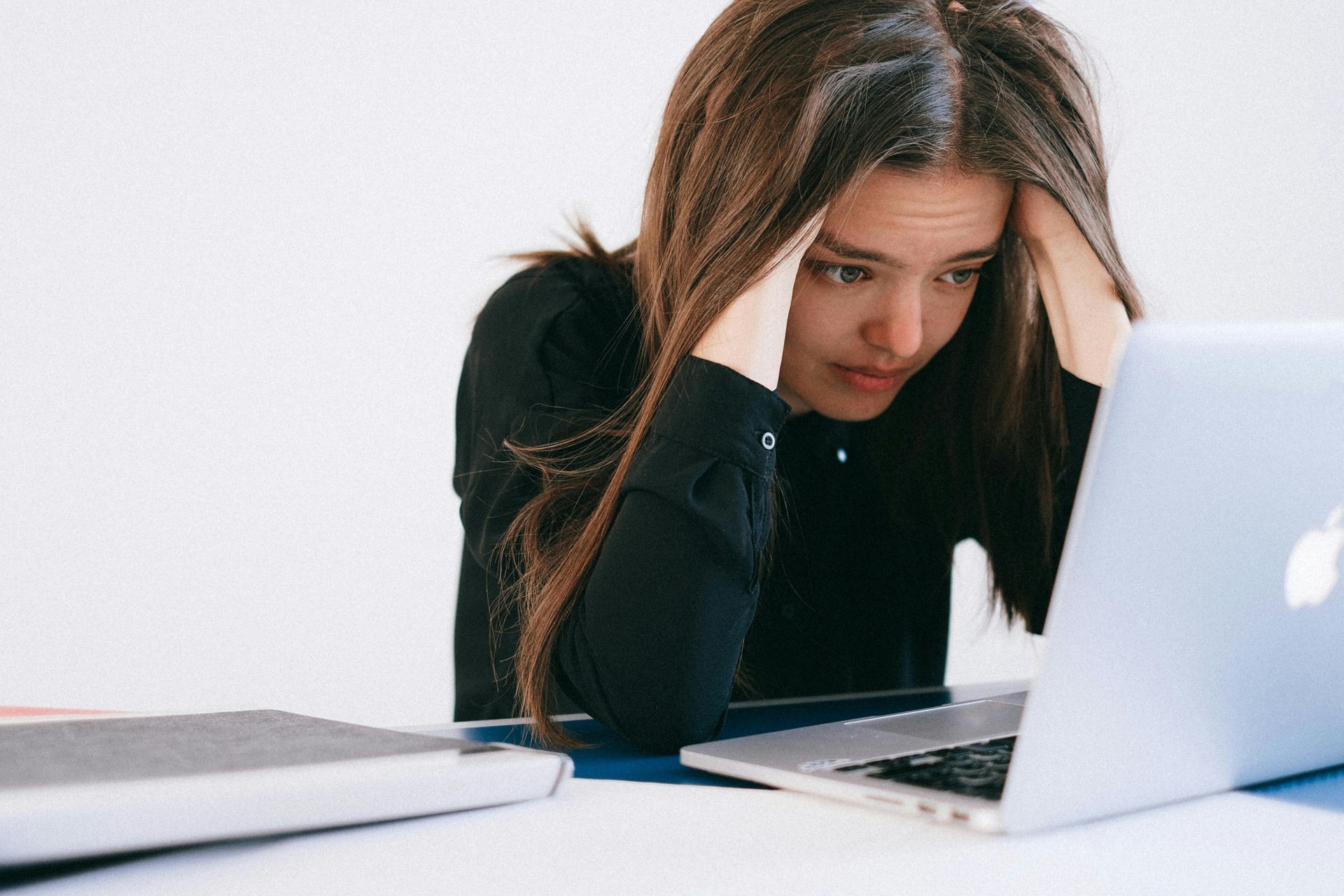 Woman with hands on head looking distressed at a laptop on a white desk.