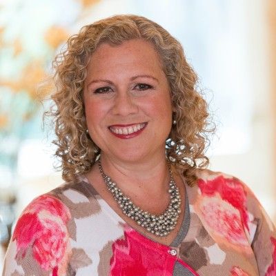 A woman with curly hair is wearing a pink floral sweater and a pearl necklace.