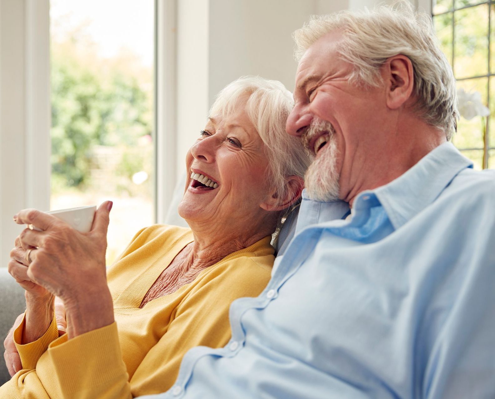 An elderly couple is sitting on a couch laughing and drinking coffee.