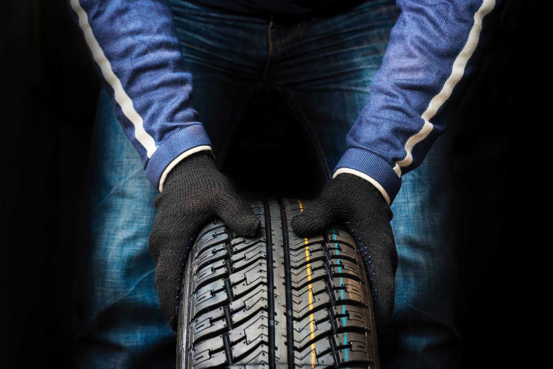 Person in jeans and blue jacket, black gloves, holding a tire against a black background. Person in jeans and blue jacket, black gloves, holding a tire against a black background.