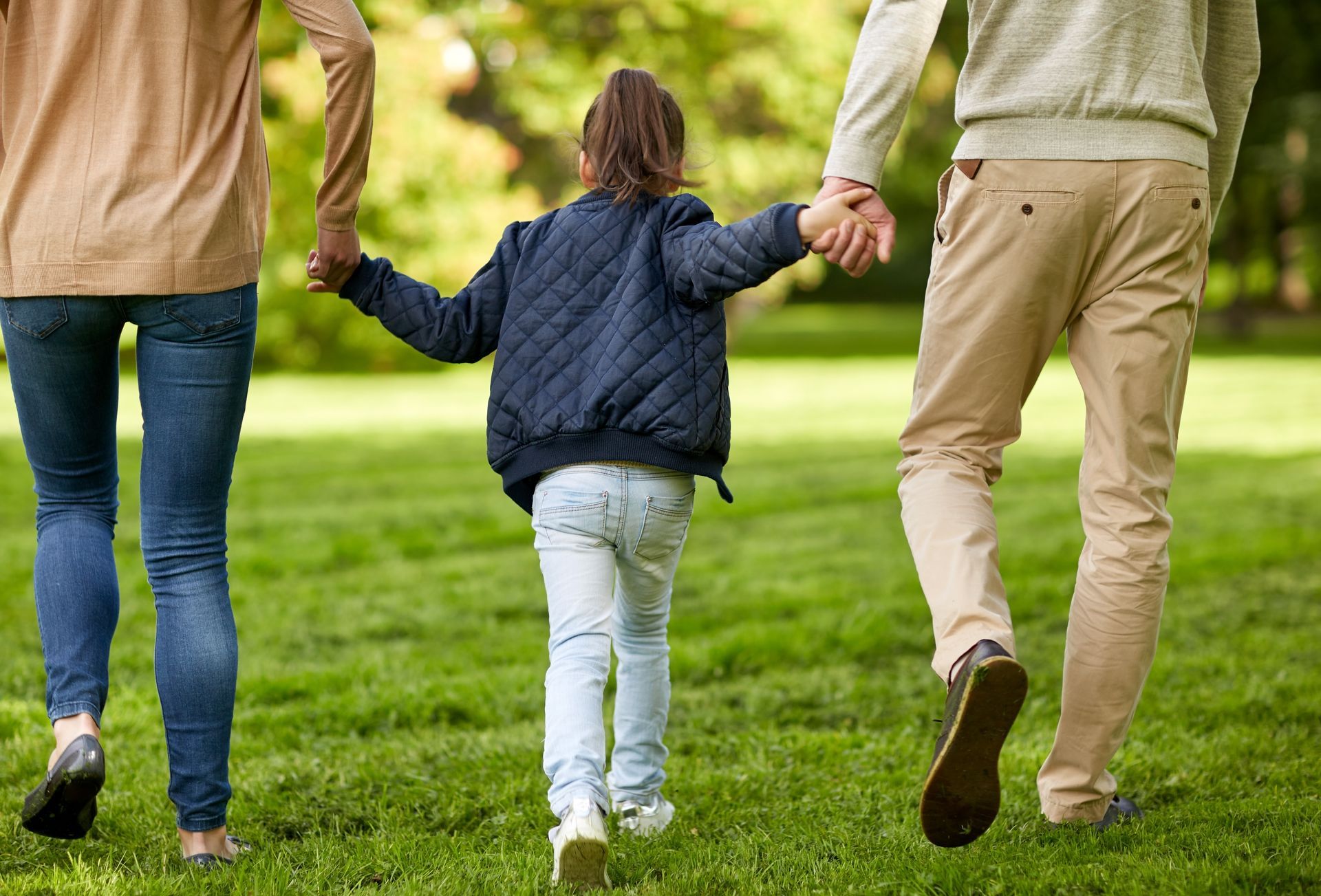 Family walking in a grassy area, holding hands; mother on left in jeans, father on right in khaki pants, child in the middle.