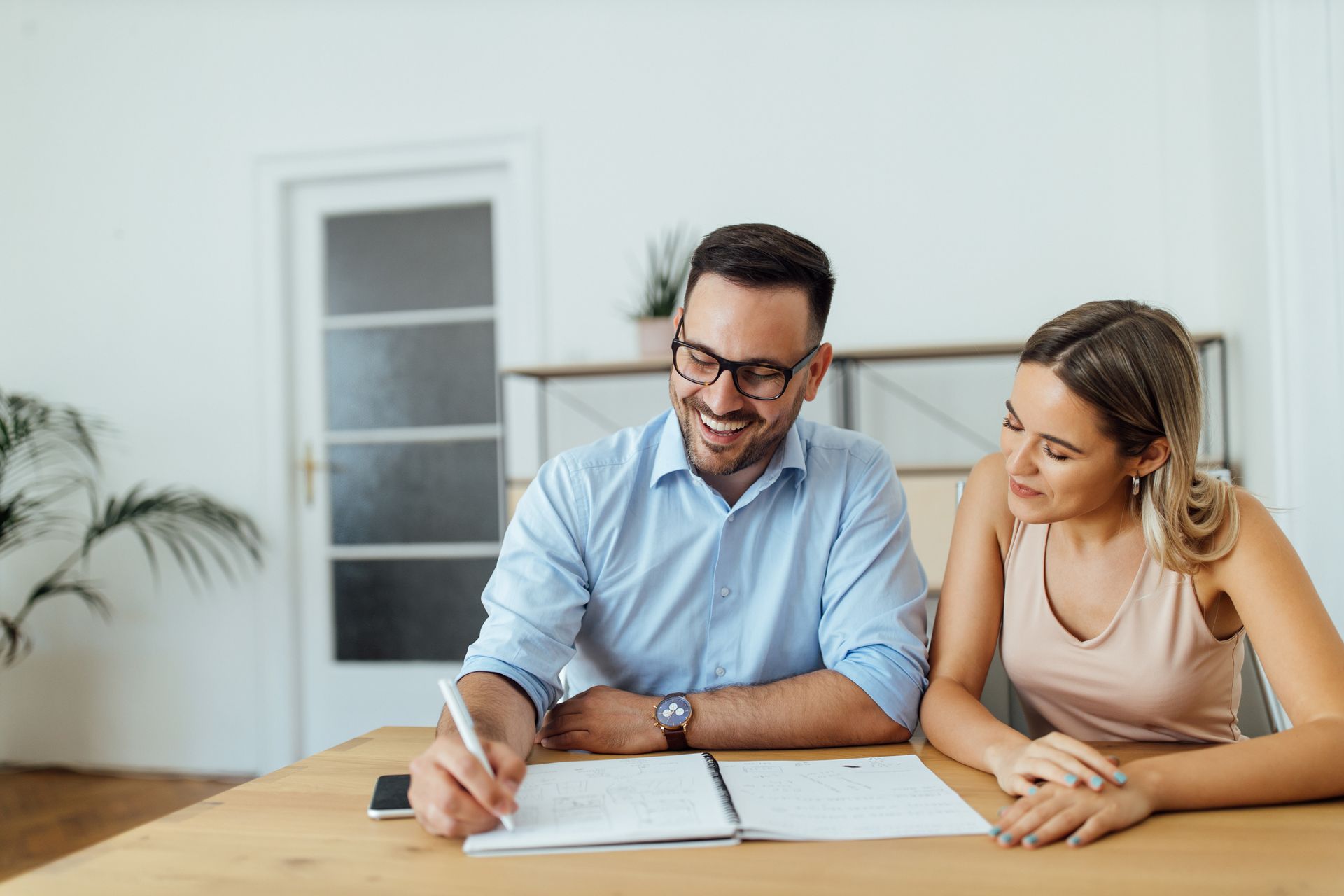 Man in glasses smiles, writes in a notebook as woman looks on; wooden table, indoor setting.
