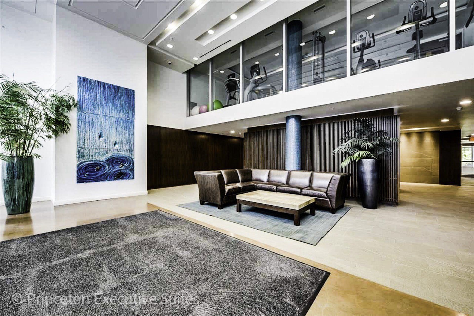 Large marble lobby area stunningly decorated with giant tropical plants and modern blue abstract wall artwork.   Large brown leather sectional couch sits in the centre of the lobby.   A rectangle brown and beige coffee  table sits in front of the sectional.    Above the sectional is a large window overlooking the lobby inside you can see fitness training equipment|princeton suites edmonton furnished suites