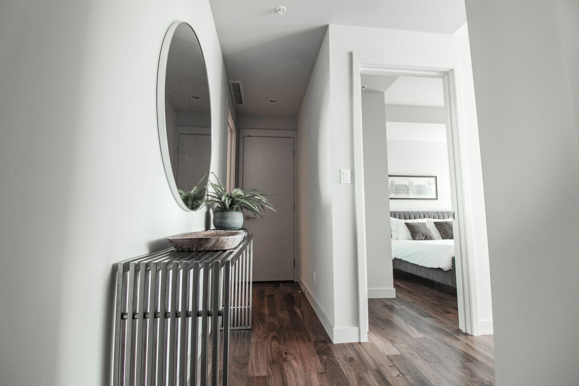 Hallway with a metal console table, round mirror, and open doorway to a bedroom with a bed.