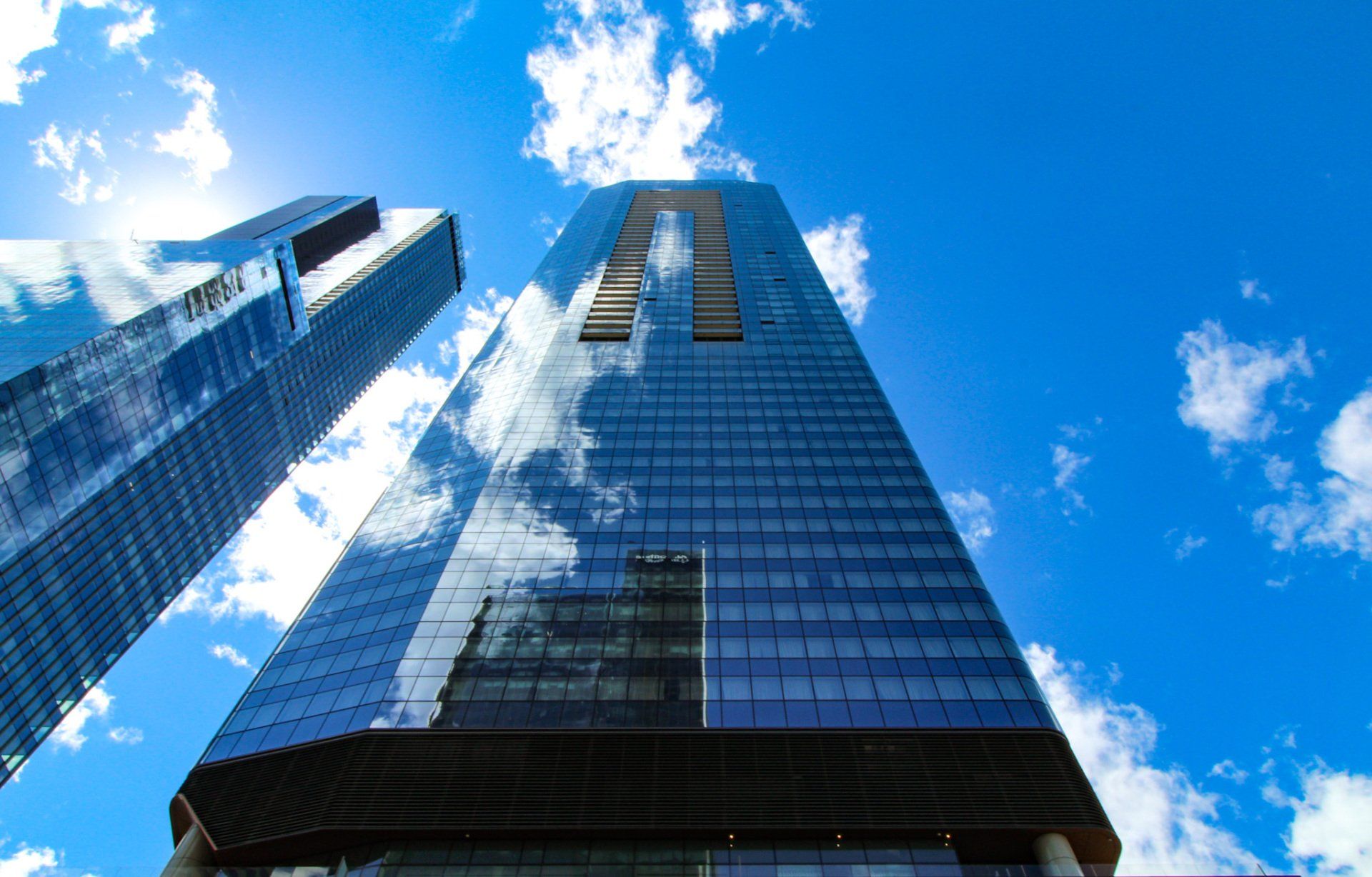 Two glass skyscrapers against a bright blue sky with clouds; sun reflects on the buildings.