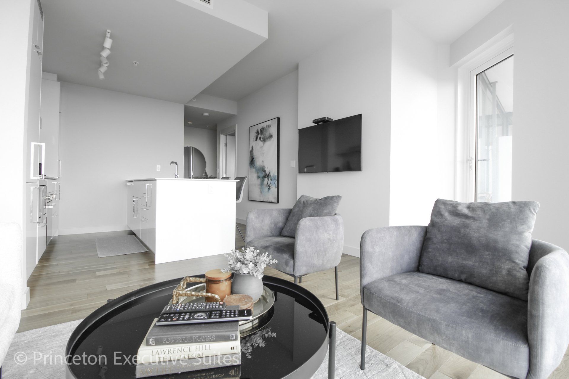 Luxurious living area in this edmonton furnished apartment with white modern kitchen in the back ground.   Grey fabrics accent chairs  in front round smoked glass coffee table.  A large black Led Tv is mounted to the wall in the back ground.