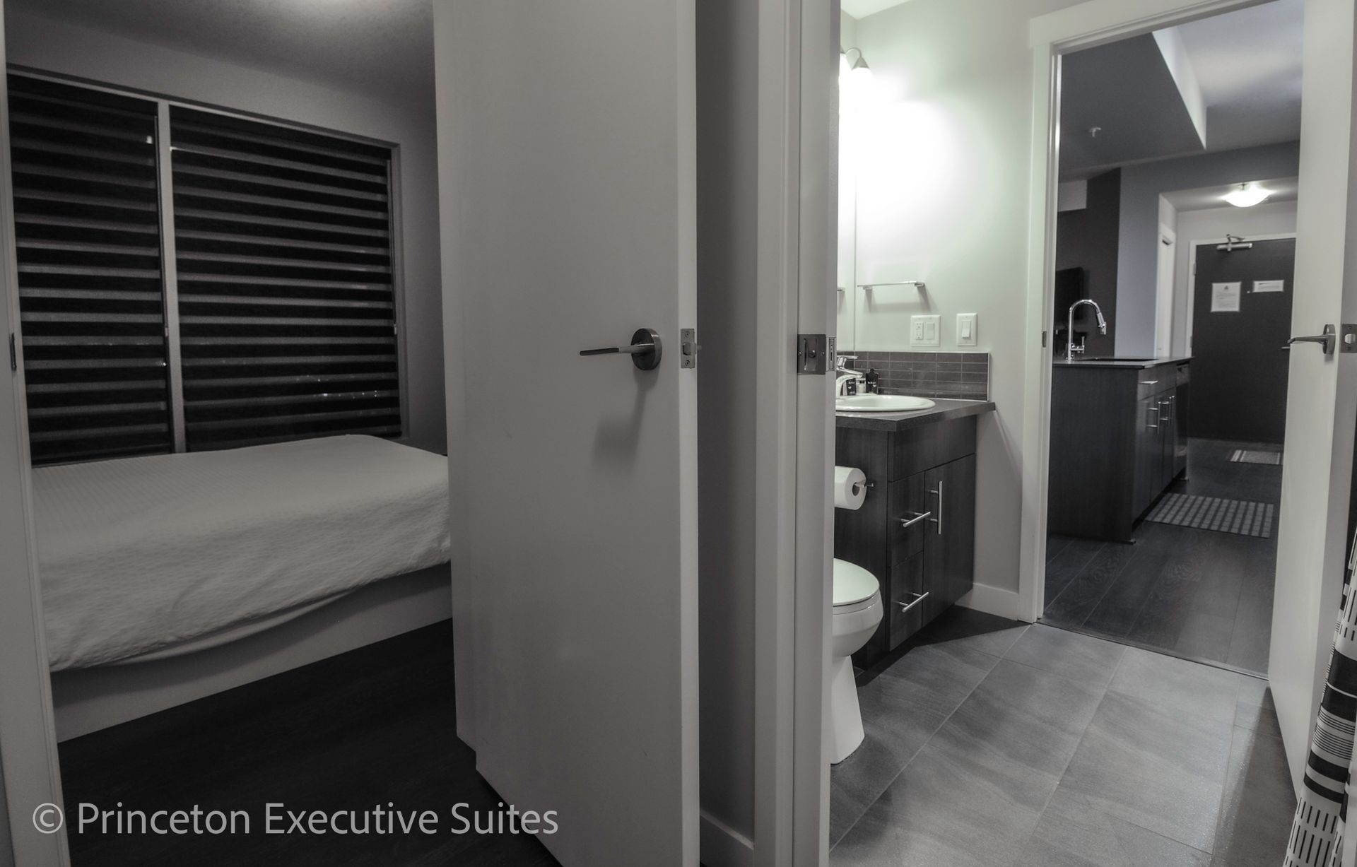 Monochrome view of a bedroom with a bed, leading to a bathroom and a hallway.