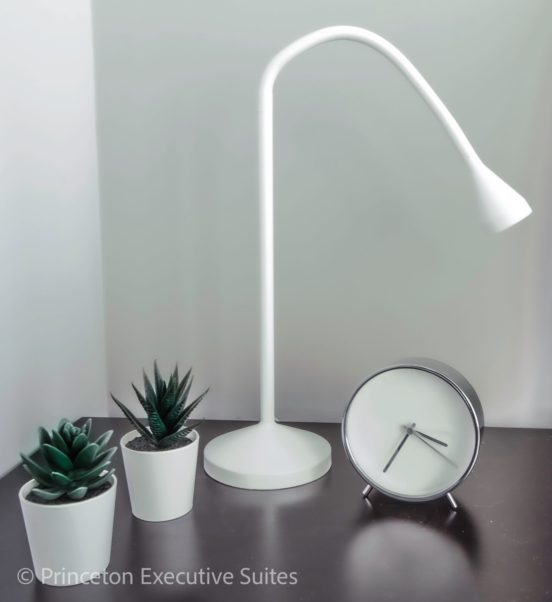 White desk lamp, clock, and potted succulents on a dark surface against a white wall.