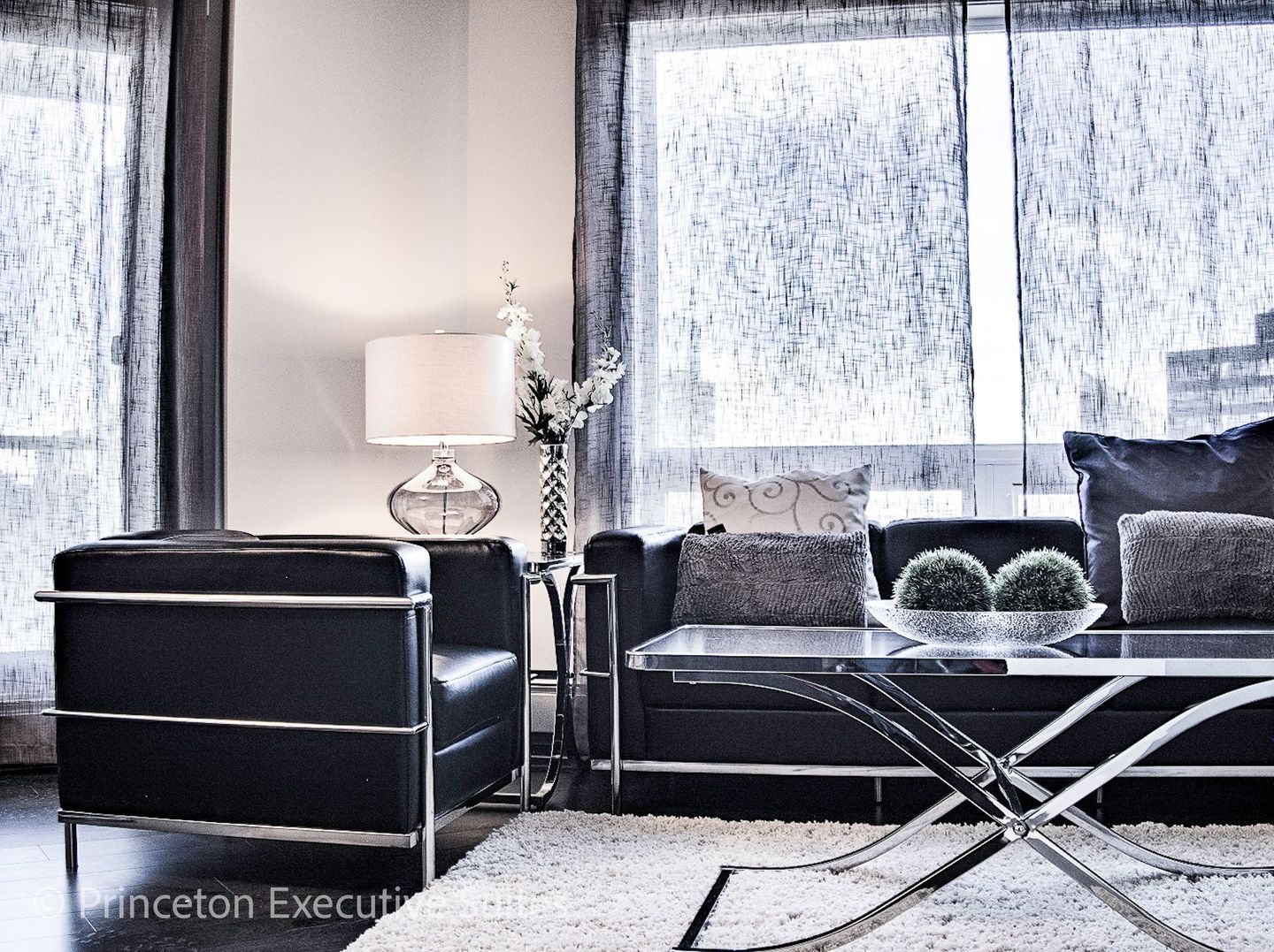 A modern furnished apartment with black leather couch chair and chair with  chrome metal and glass coffee table in front of it. 