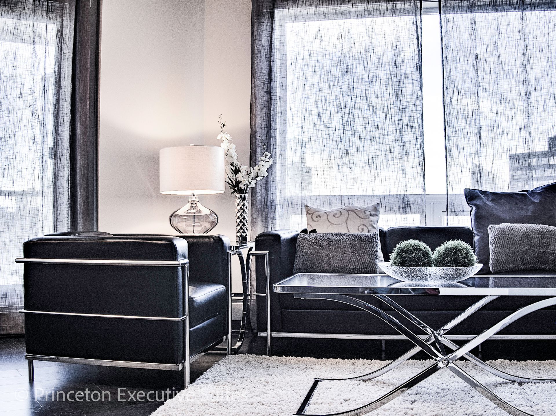 A modern furnished apartment with black leather couch chair and chair with metal and glass coffee table in front of it. 