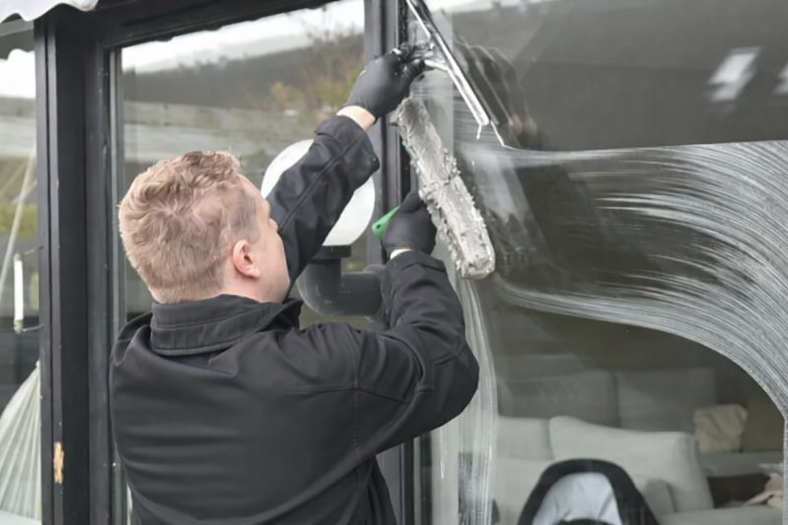 Man wearing black gloves and jacket cleans a window with a squeegee.