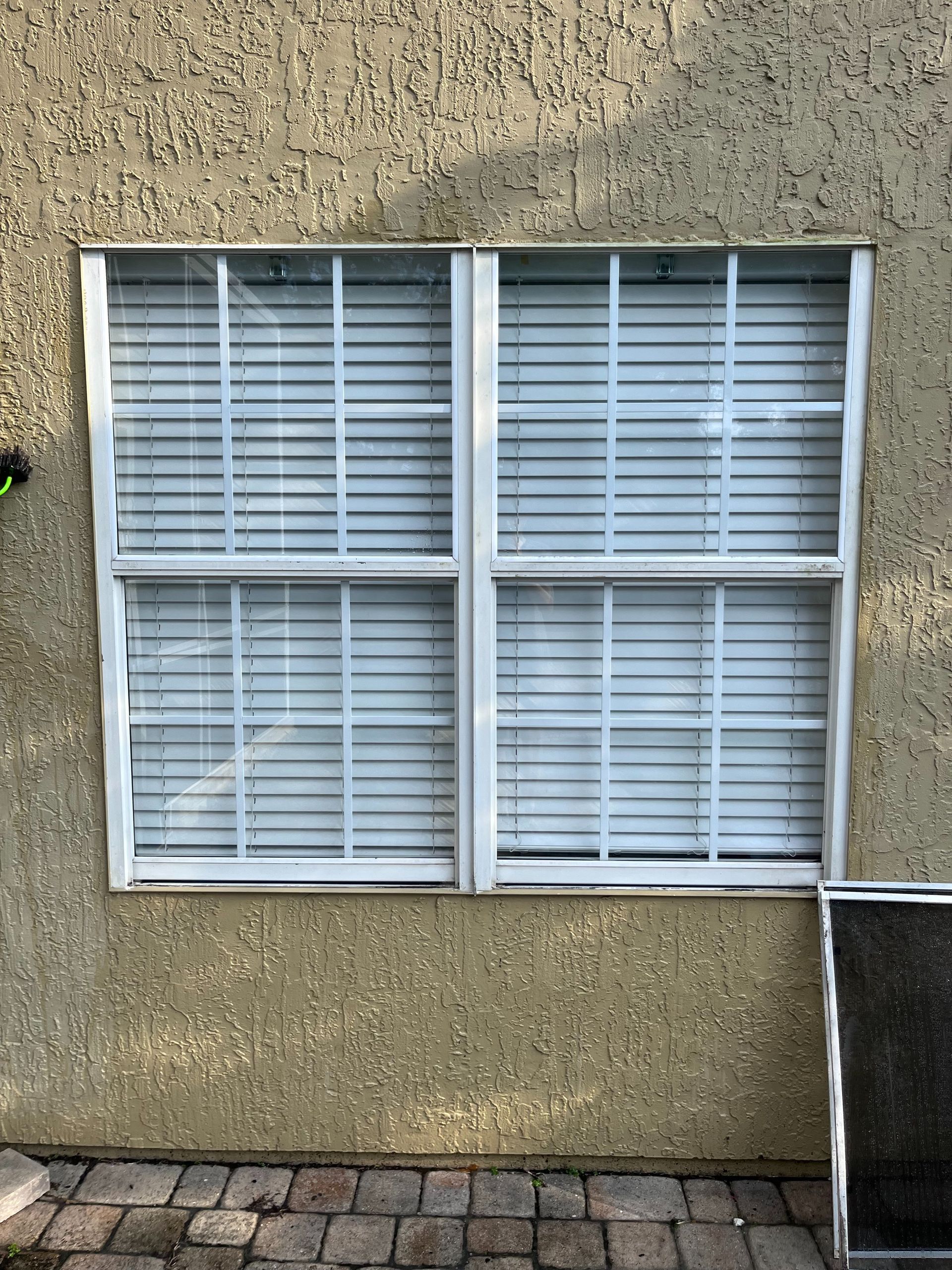 White-framed window with blinds, on a textured tan wall, above brick paving.