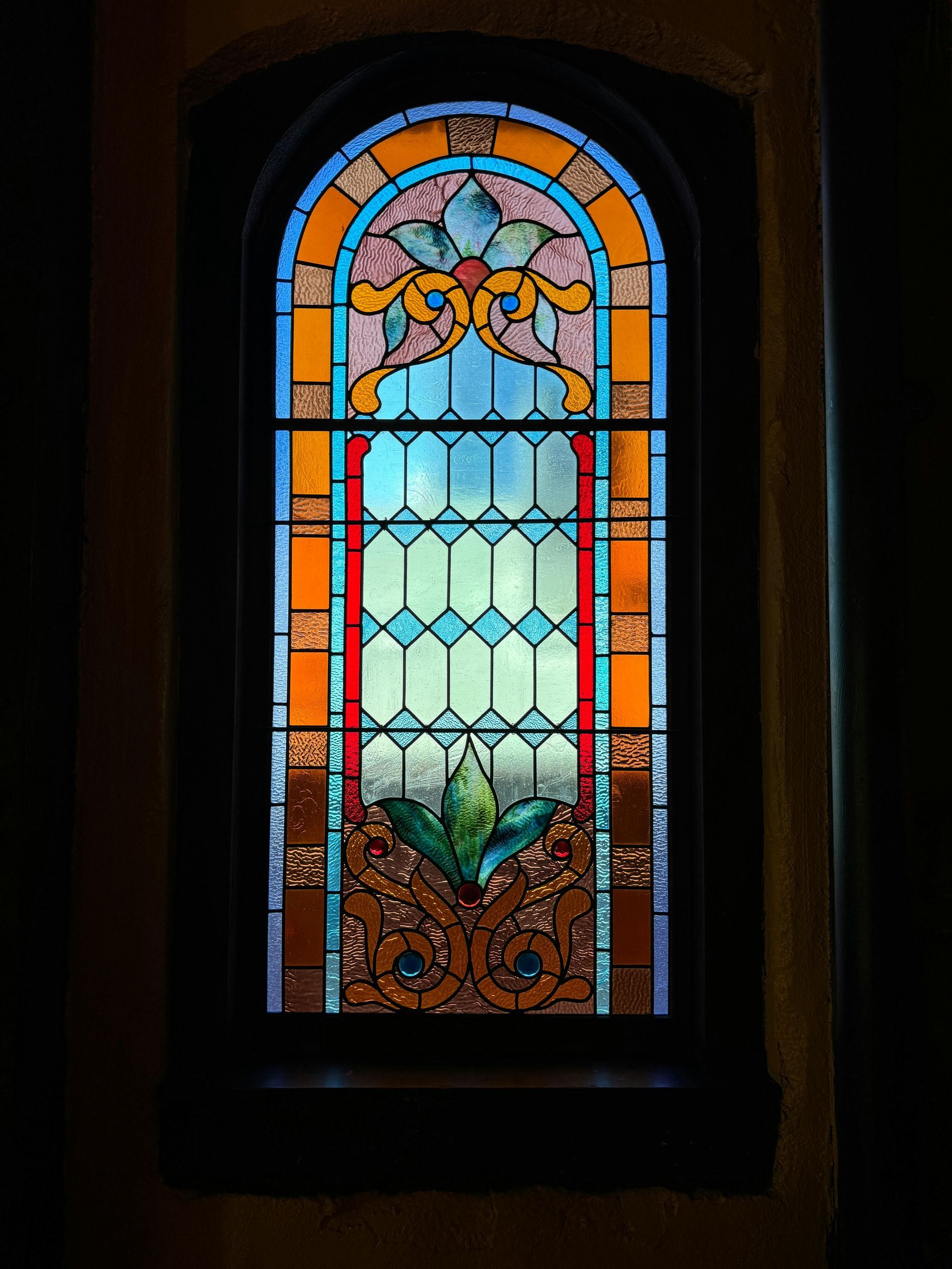 Stained glass window with an arched top, featuring floral and geometric designs, set in a dark frame.