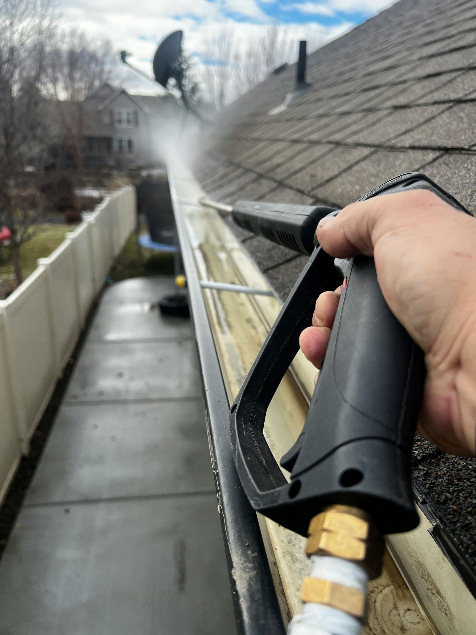A person cleaning a roof gutter with a pressure washer. Water sprays over the gutter's edge.