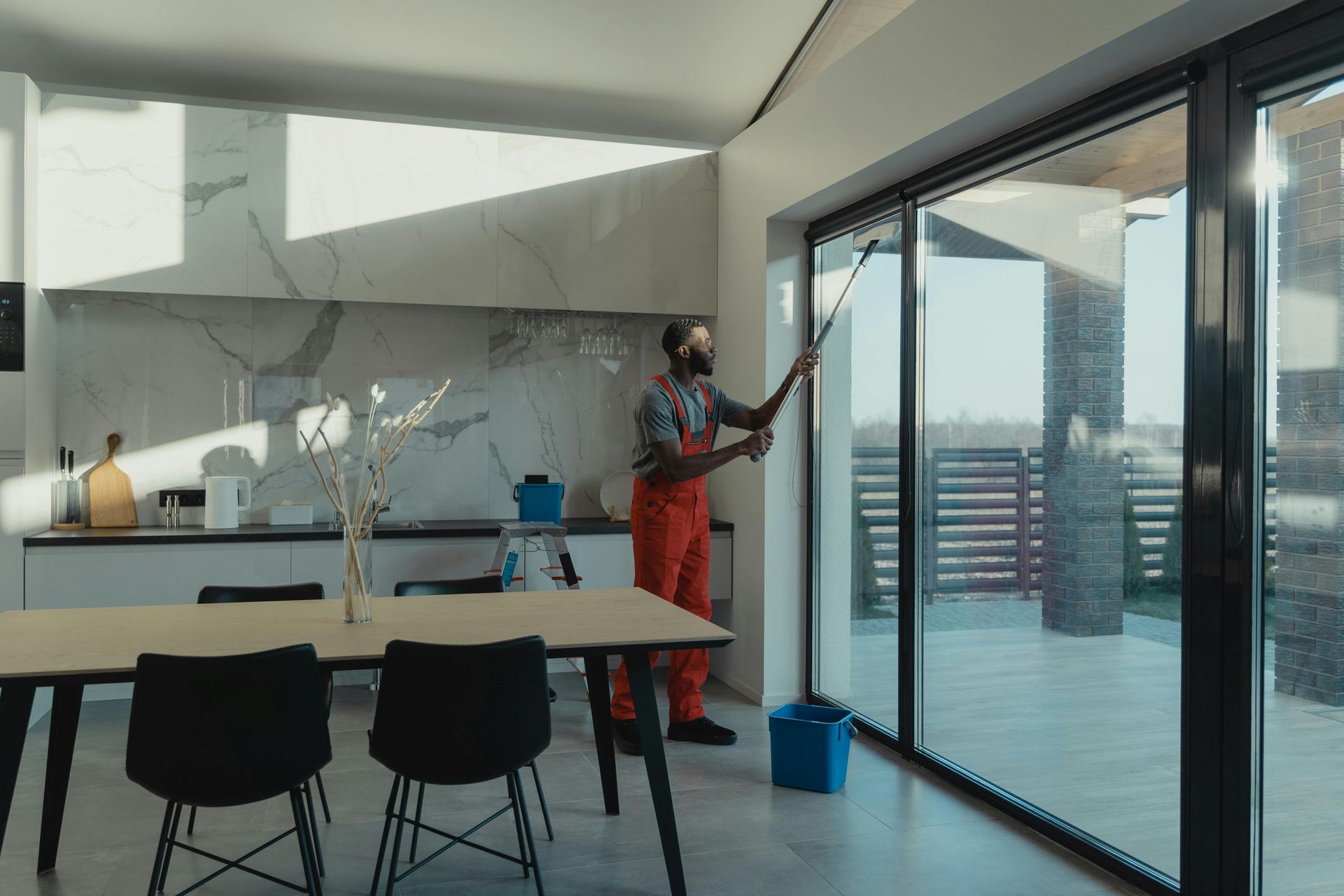 Man in orange coveralls cleaning a large window with a squeegee in a modern kitchen.