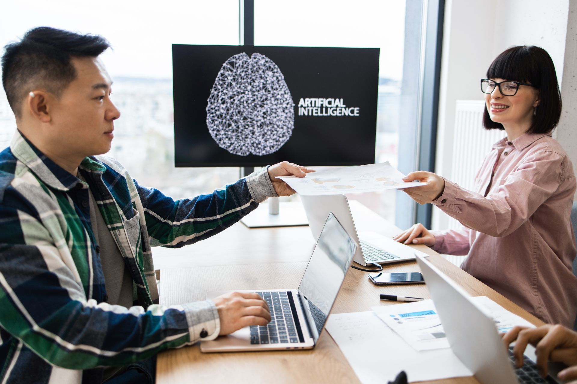 People in office, passing documents, with AI brain graphic on screen.