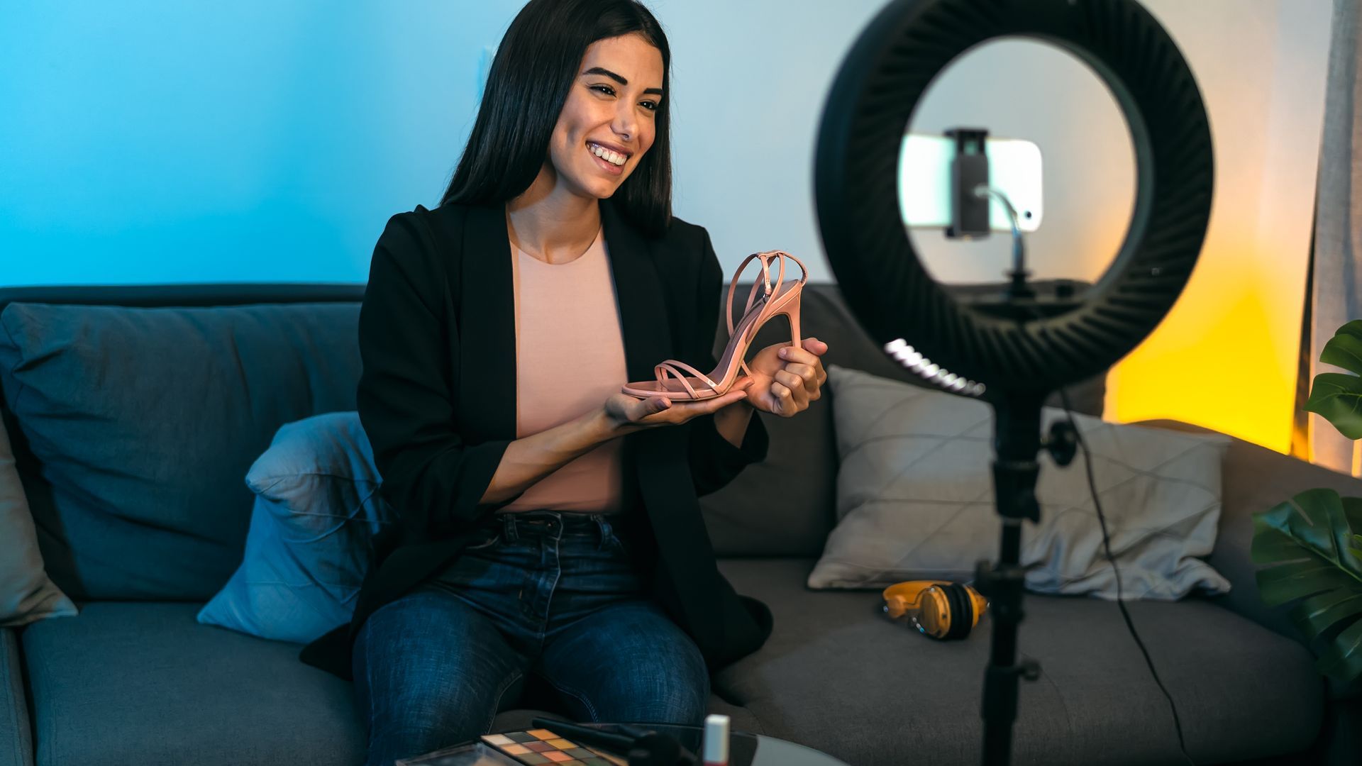 A woman is sitting on a couch holding a pair of shoes in front of a ring light.