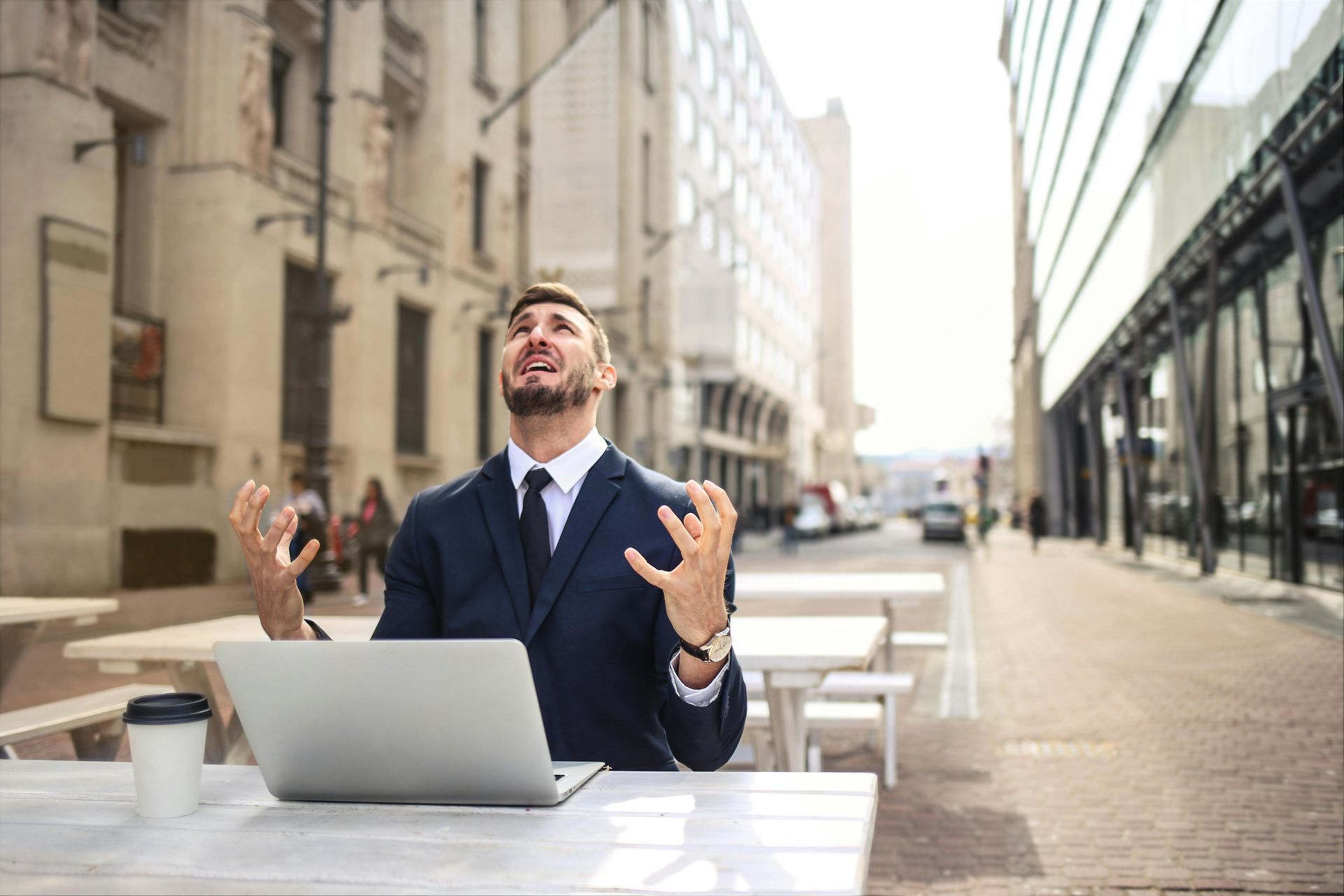Man in suit looking frustrated, arms raised, sitting at a table with a laptop and coffee in a city setting.