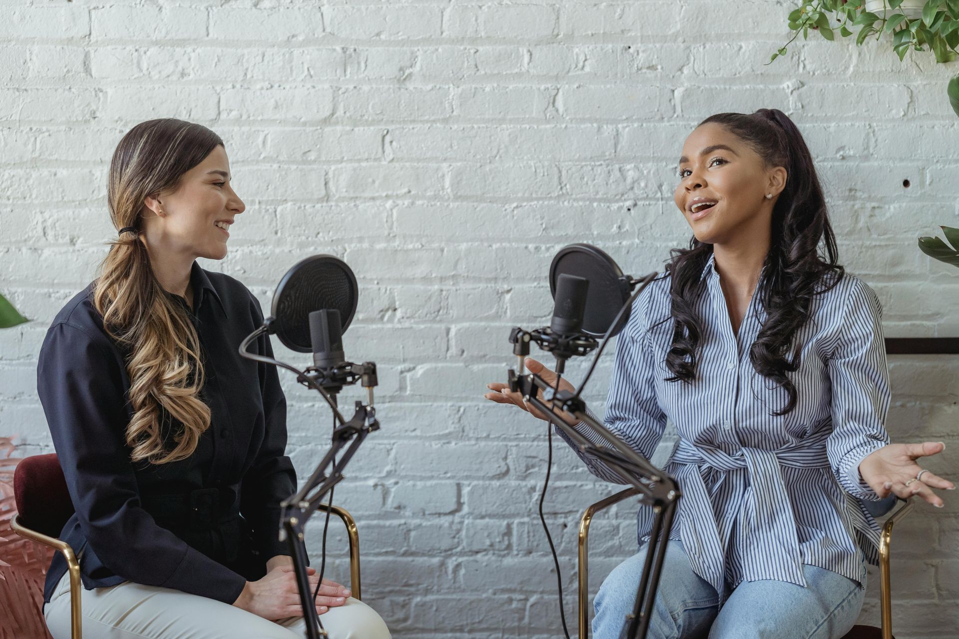 Two women recording a podcast, seated at microphones in front of a white brick wall. One gestures with hands.