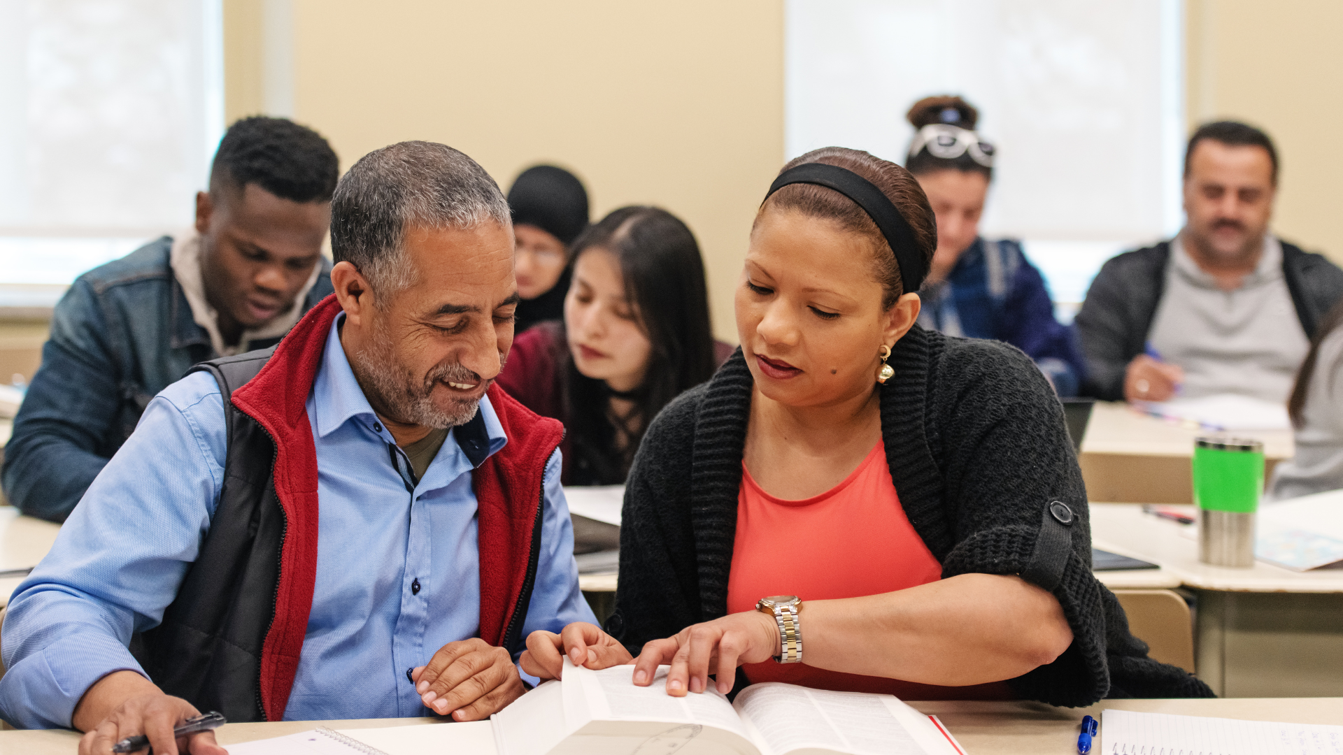 A man and a woman are sitting at a table in a classroom looking at a book.