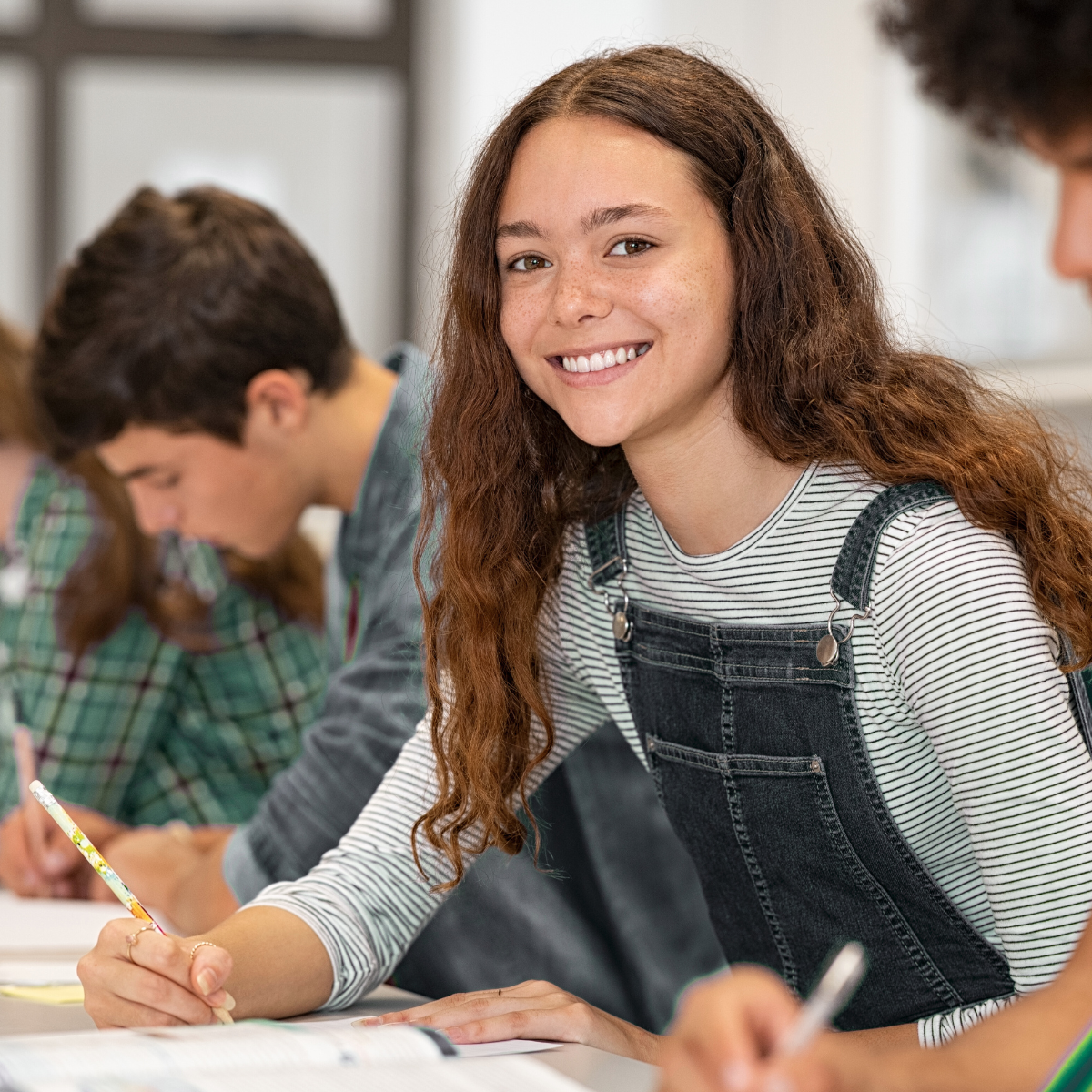A girl is smiling while sitting at a desk in a classroom with other students.