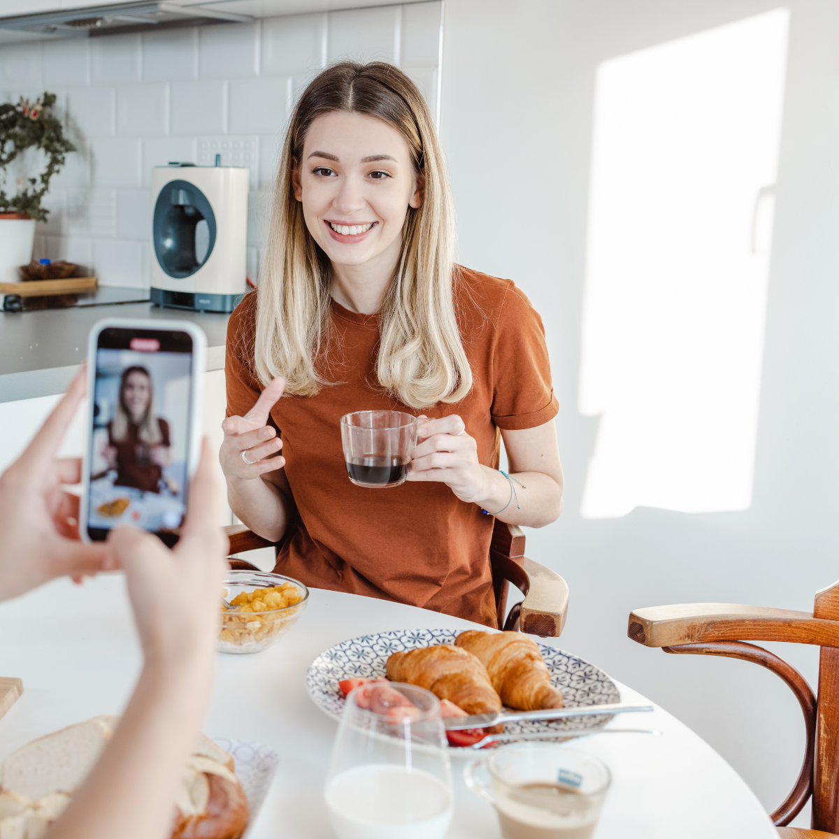 A woman is sitting at a table holding a cup of coffee while another person takes a picture of her.