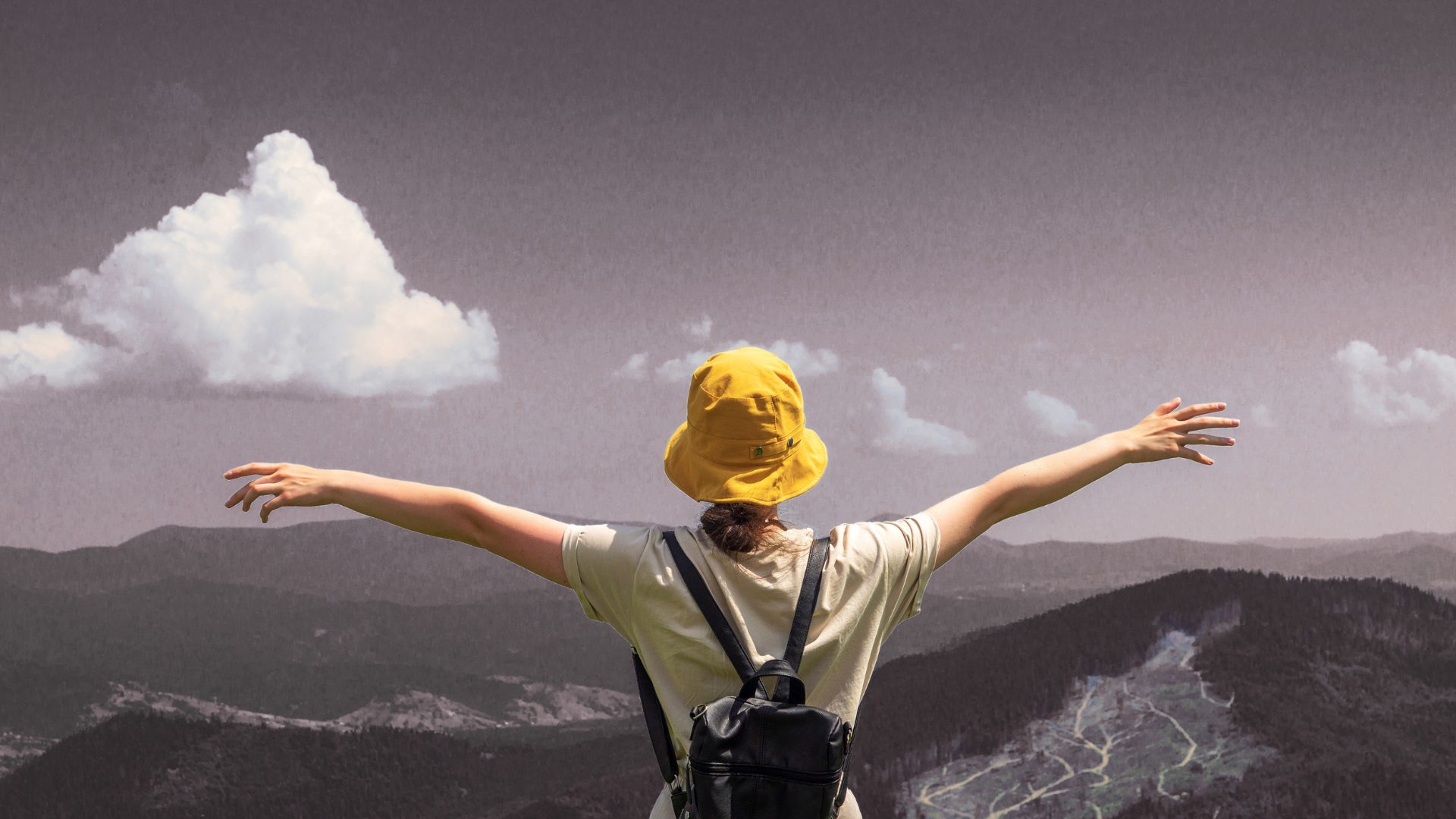 A person wearing a yellow hat and a backpack is standing on top of a mountain with their arms outstretched.