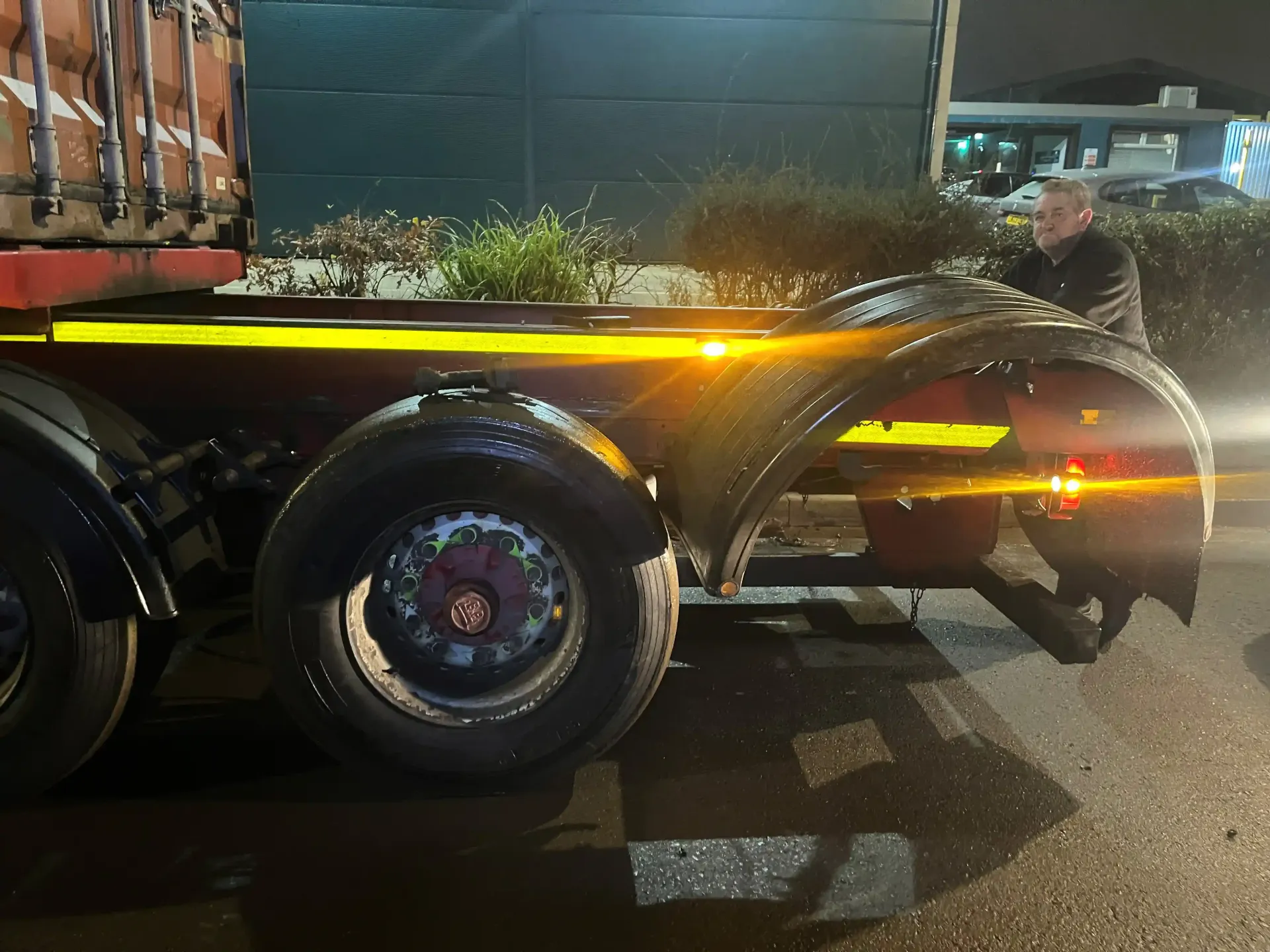 A person stands at night next to a large, dark trailer with dual wheels, glowing yellow reflective safety strips.