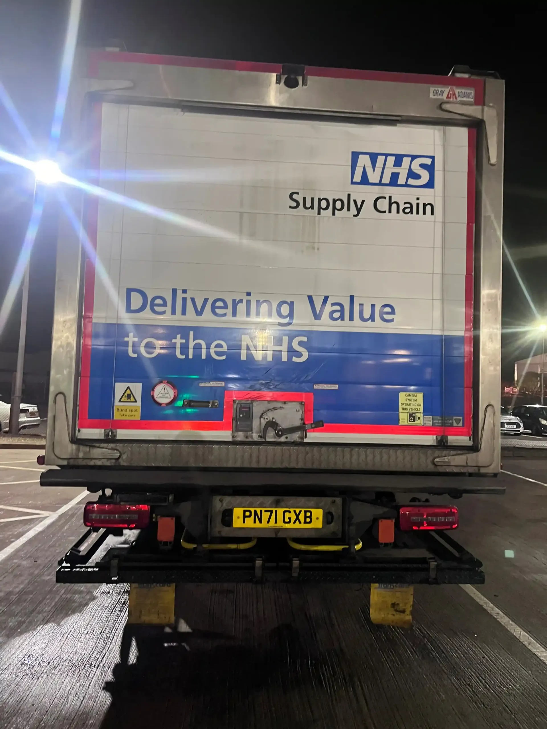 The rear of an NHS Supply Chain delivery truck parked at night, displaying the company logo and slogan.