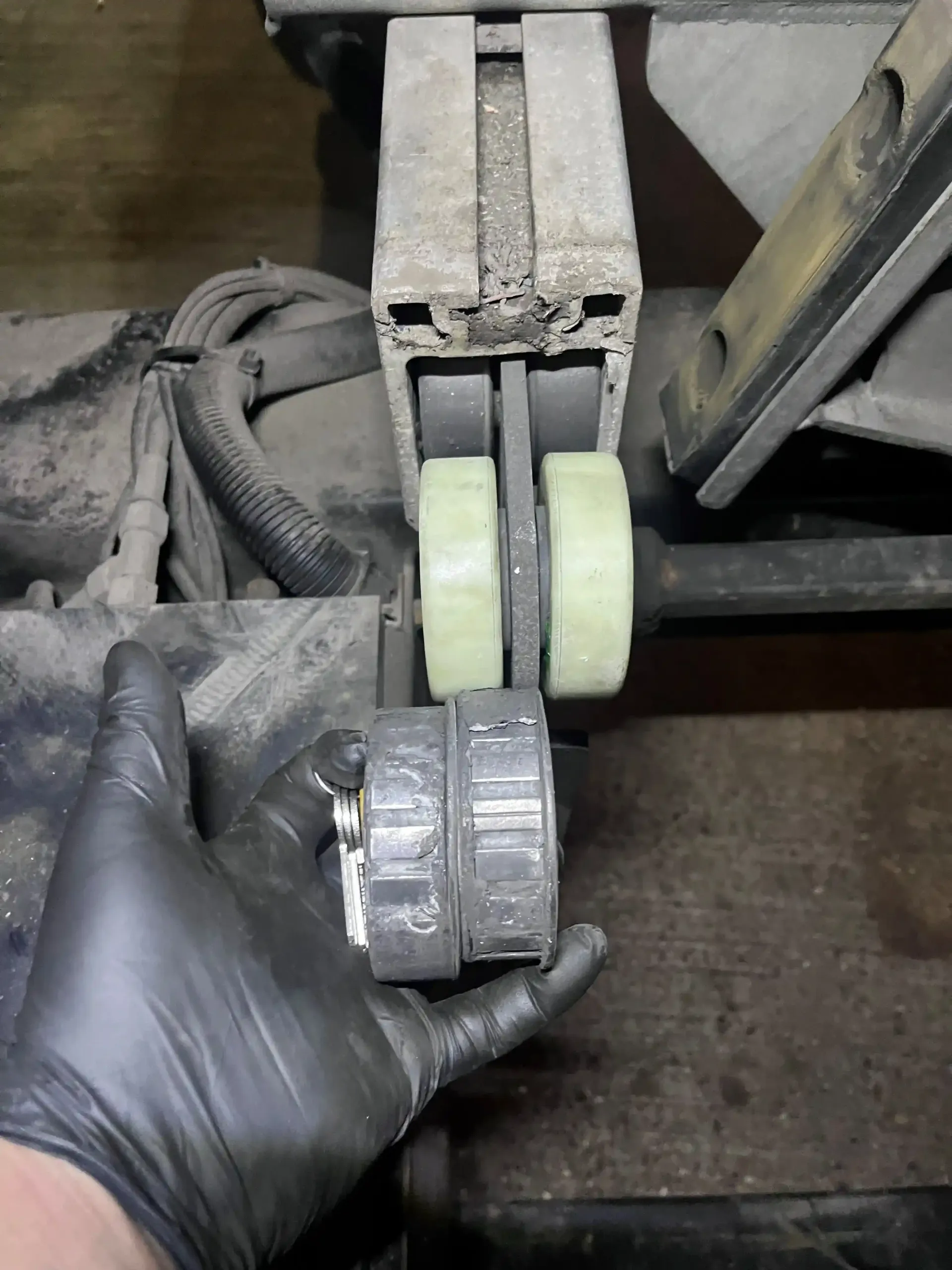 A gloved hand holds a worn, cylindrical bushing next to a mechanical assembly featuring two pale green rollers on a track.