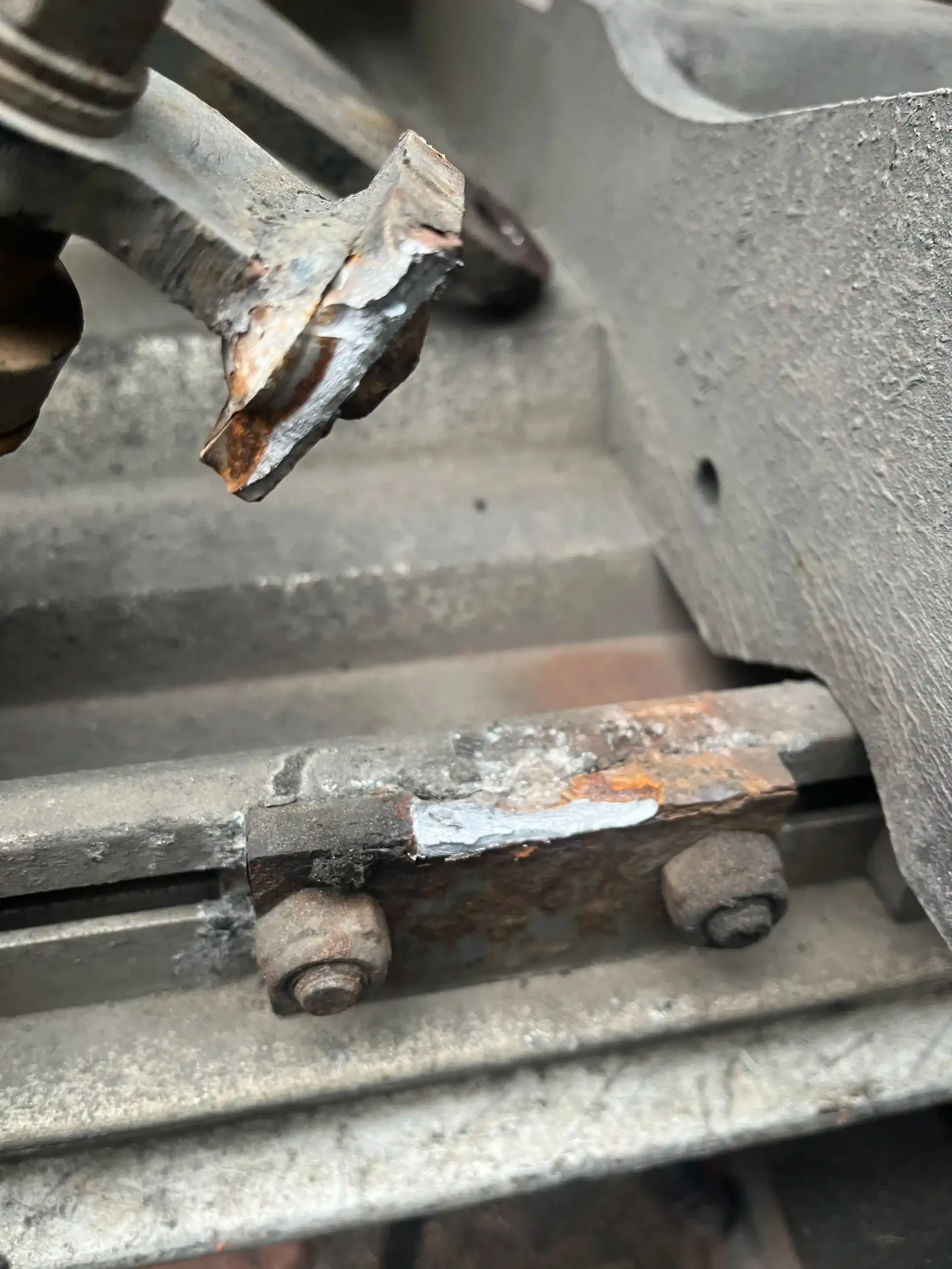 A hand holds a broken metal fitting above a rusty rail bracket with two bolts on a weathered industrial structure.