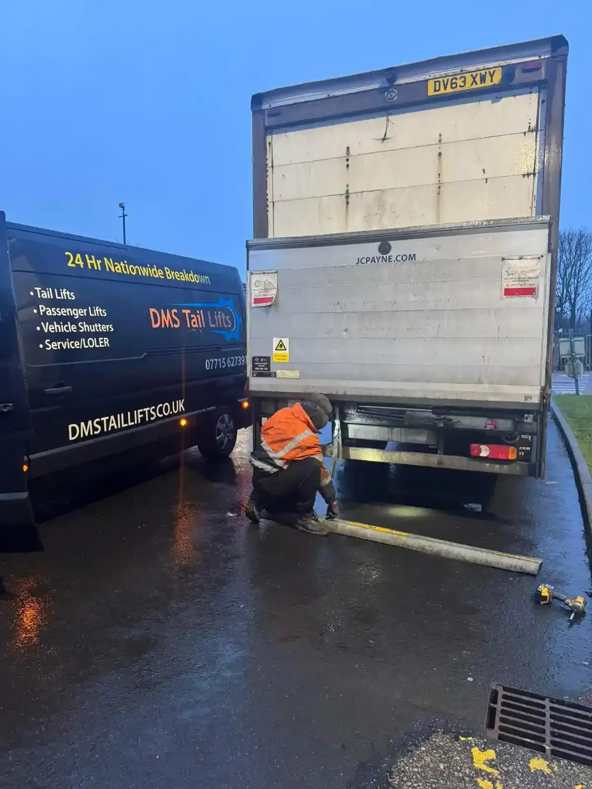 A technician in high-visibility gear works on the tail lift of a parked delivery truck during a rainy, dim day.