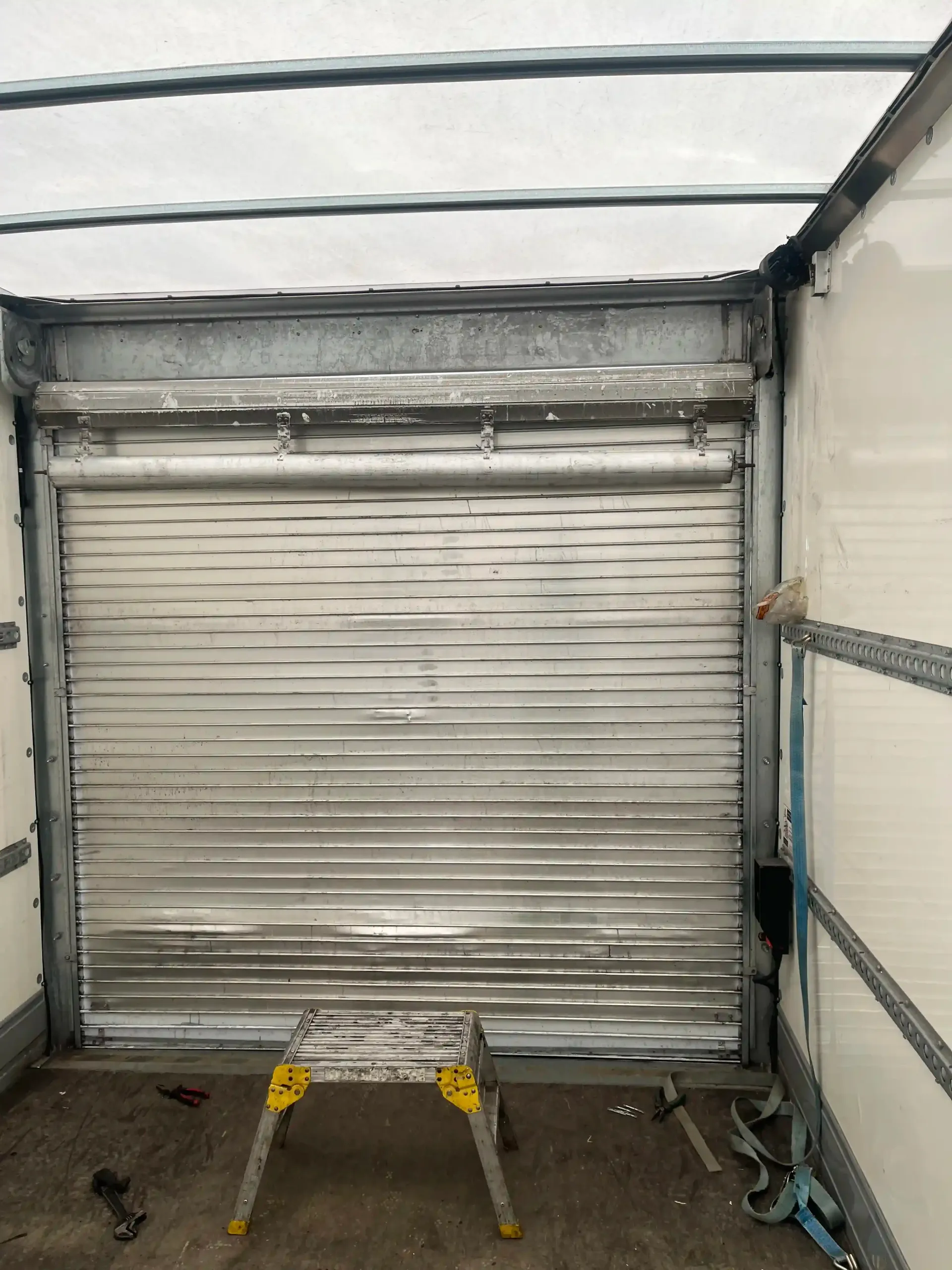 Inside a box truck trailer, a metal roll-up door is closed, with a small yellow and metal stepladder on the floor.