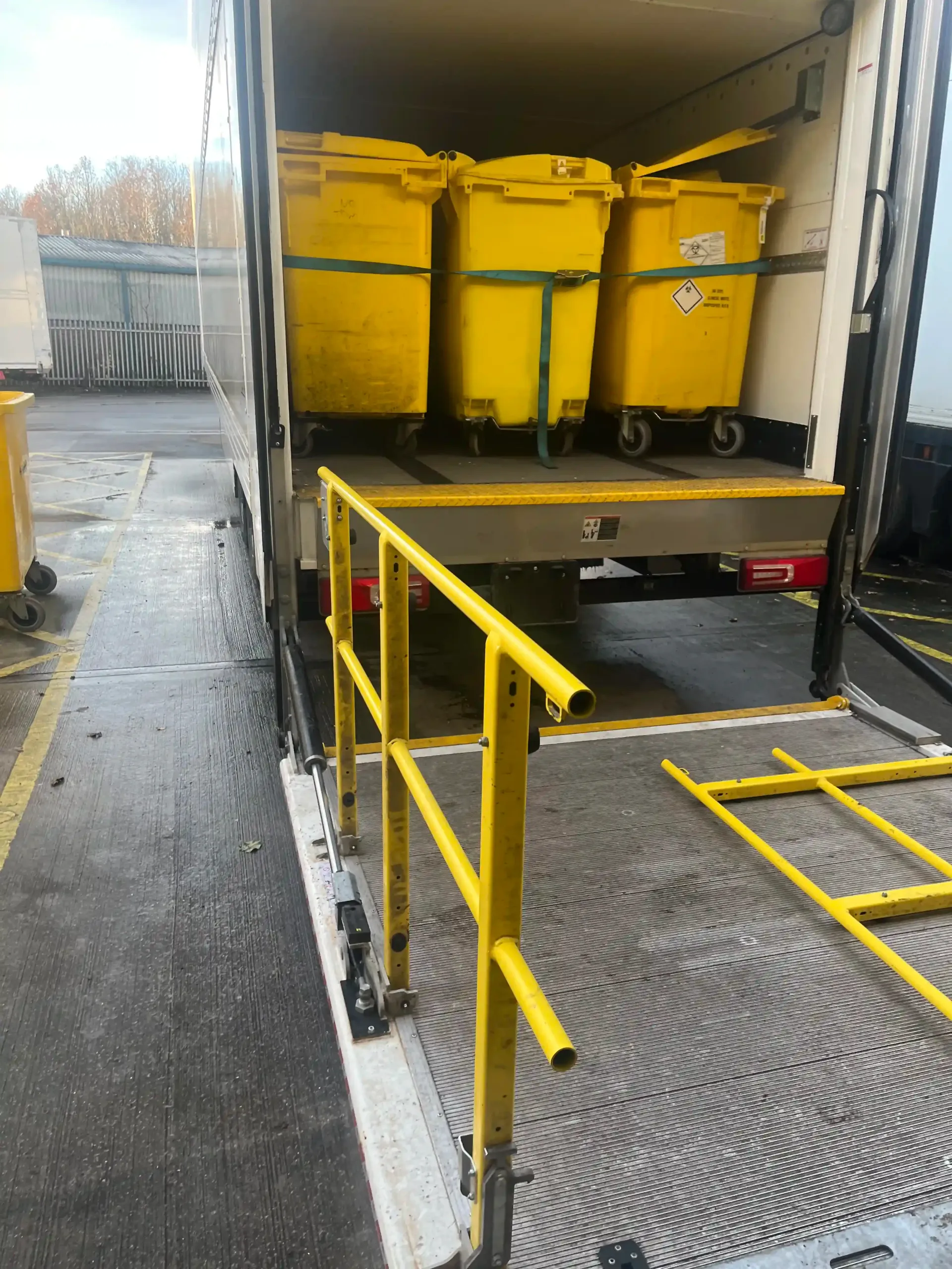 A truck interior featuring three yellow waste containers, with a yellow safety railing on the loading ramp.
