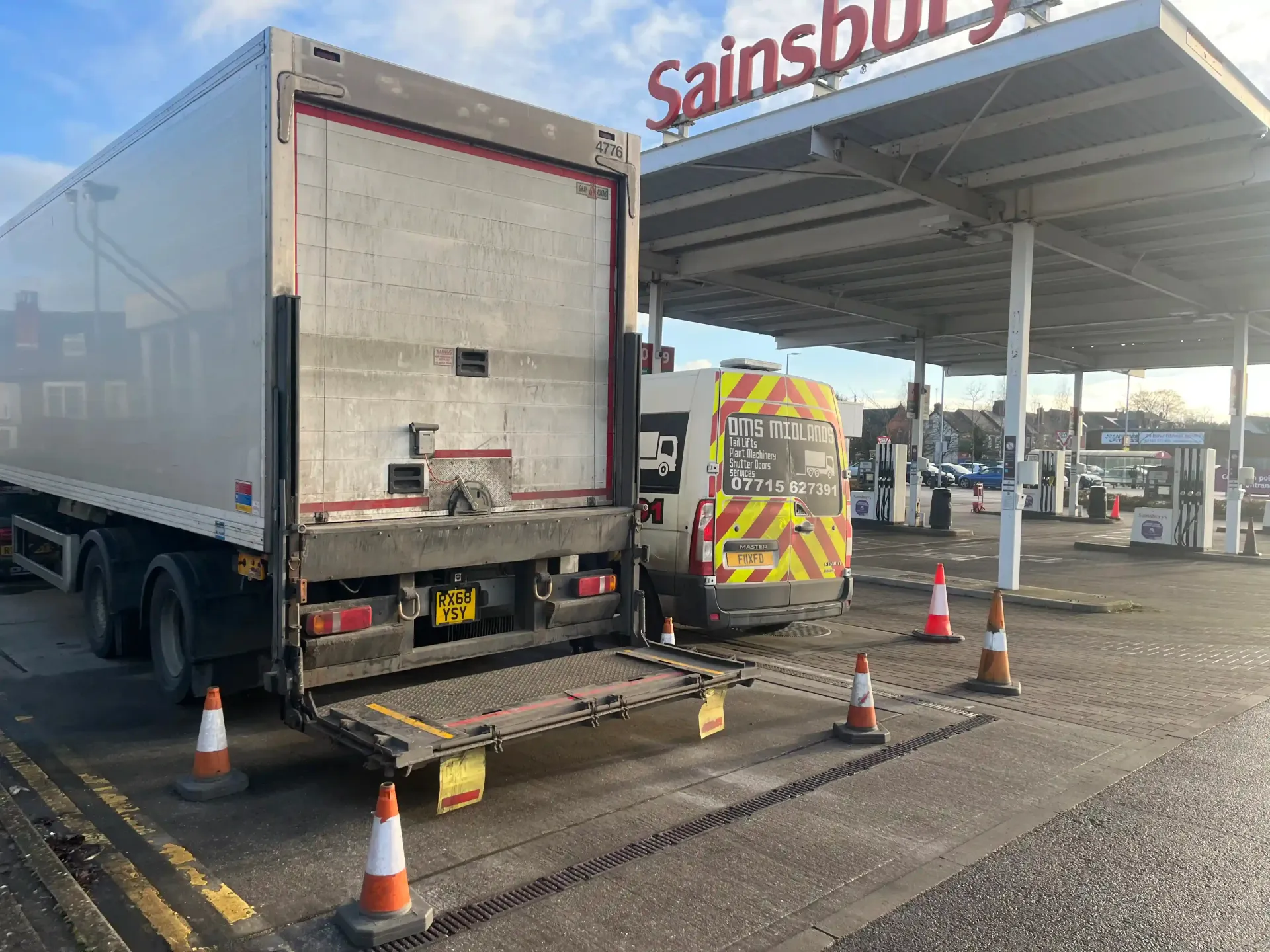 A Sainsbury’s truck with its tail lift lowered at a fuel station, surrounded by orange traffic safety cones.