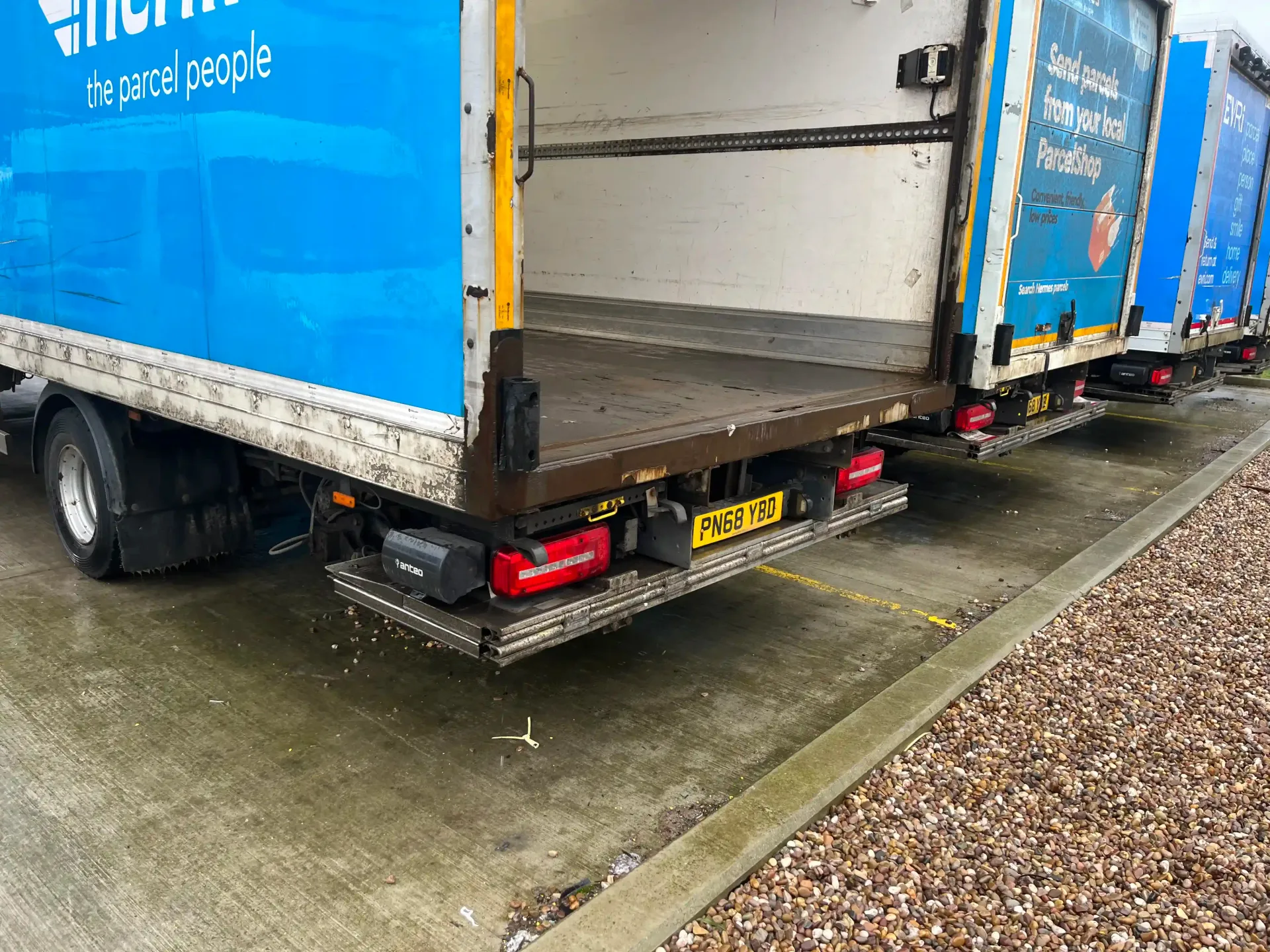 The rear of a blue and white delivery truck parked on pavement with its cargo door open and metal tail lift lowered.