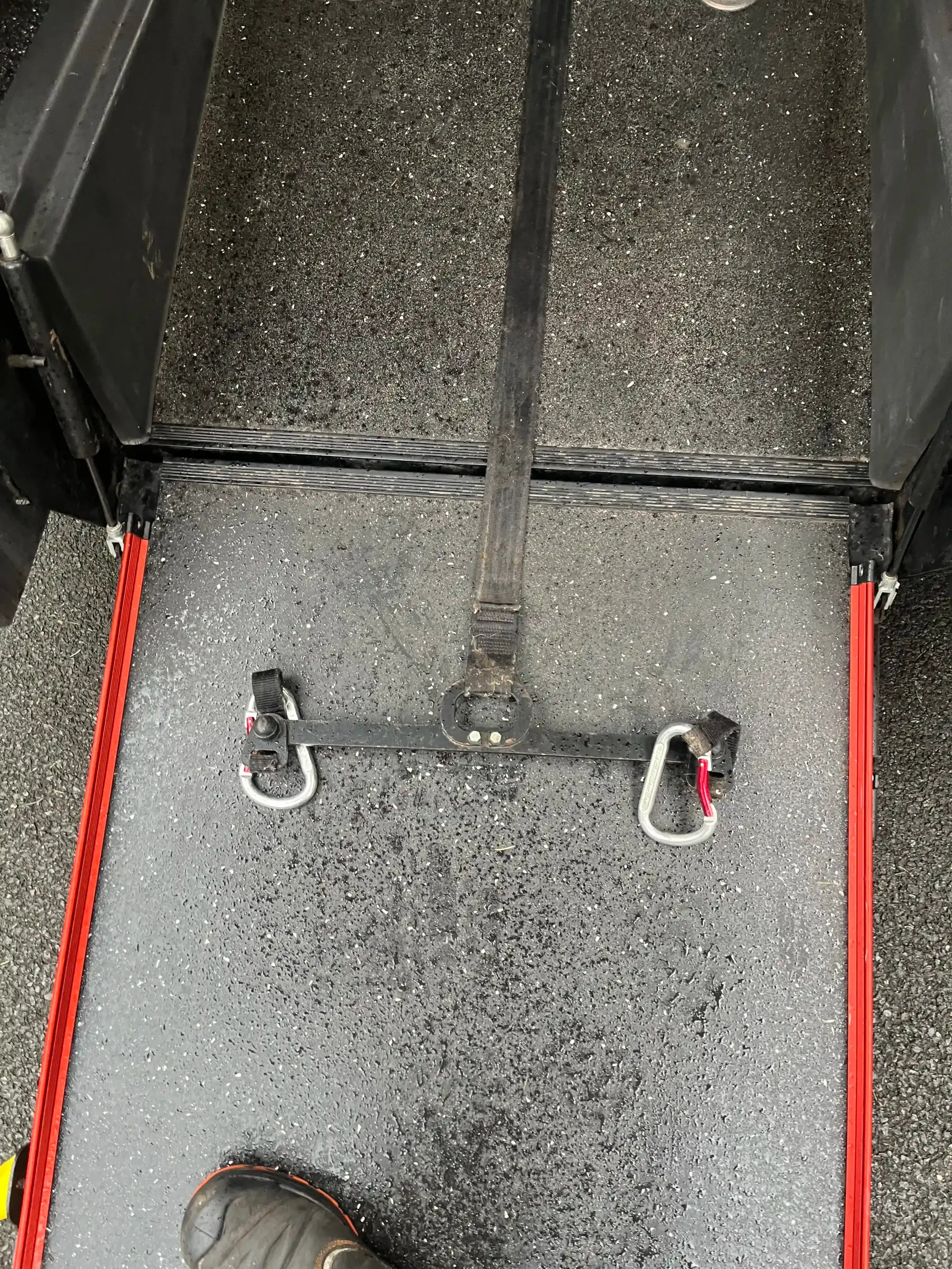 A close-up view of a wheelchair securement strap mechanism featuring two carabiners on a ramp with red side rails.