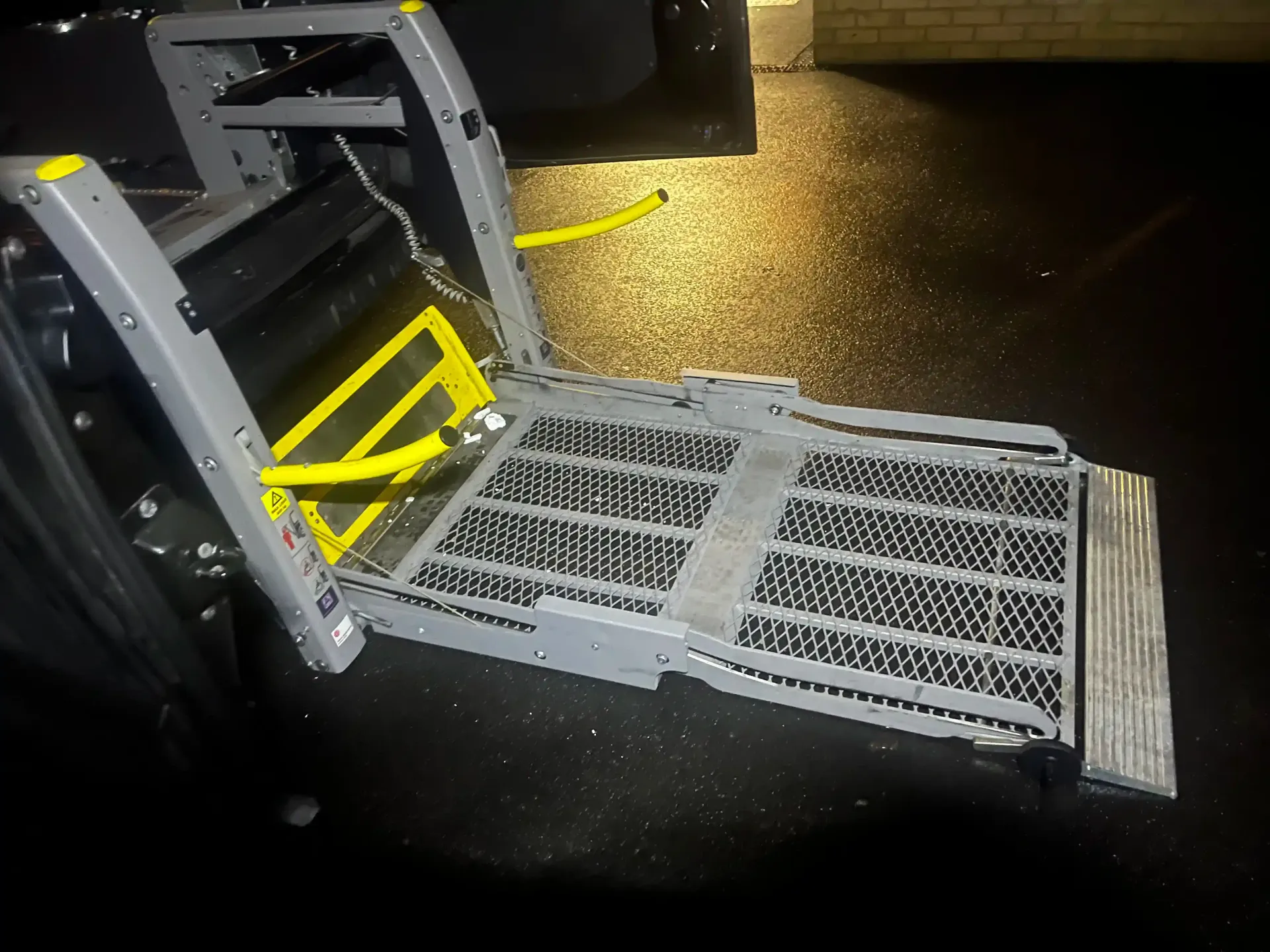 A gray vehicle wheelchair lift deployed at ground level, featuring a black mesh platform and yellow safety barriers.